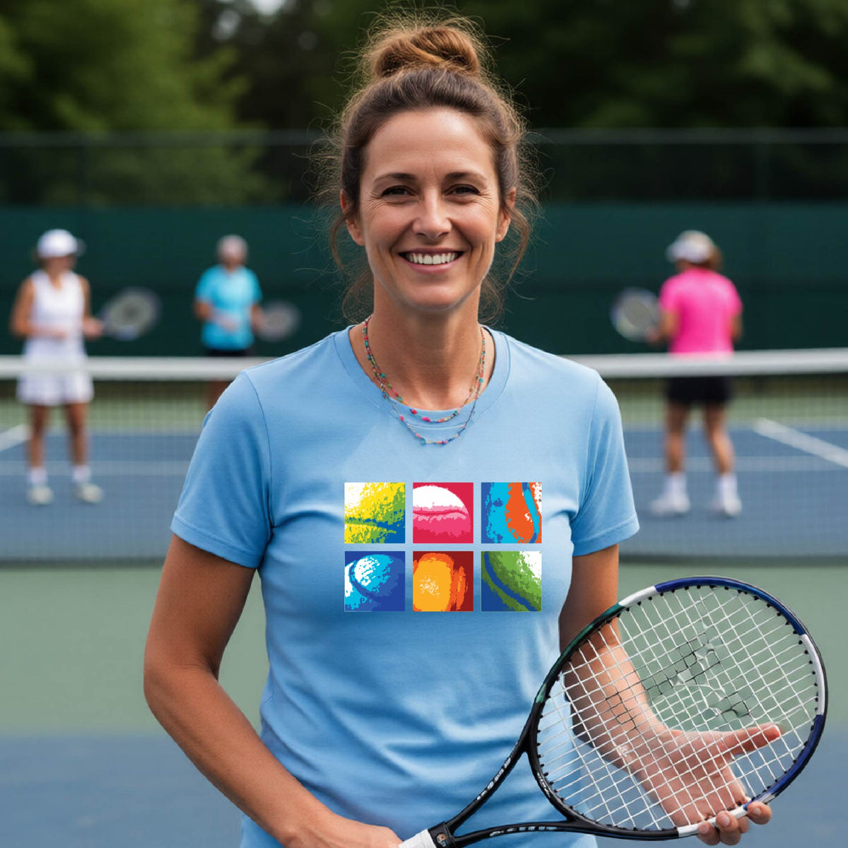 A smiling woman on a blue tennis court holds a tennis racket and wears a blue short-sleeve T-shirt with a colorful 2x3 grid graphic on the chest of tennis balls. Other tennis players are blurred in the background.