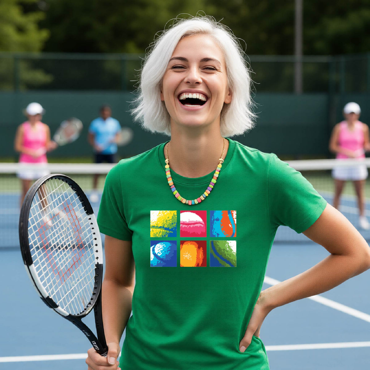 A smiling woman on a blue tennis court holds a tennis racket and wears a green short-sleeve T-shirt with a colorful 2x3 grid graphic on the chest of tennis balls. Other tennis players are blurred in the background.