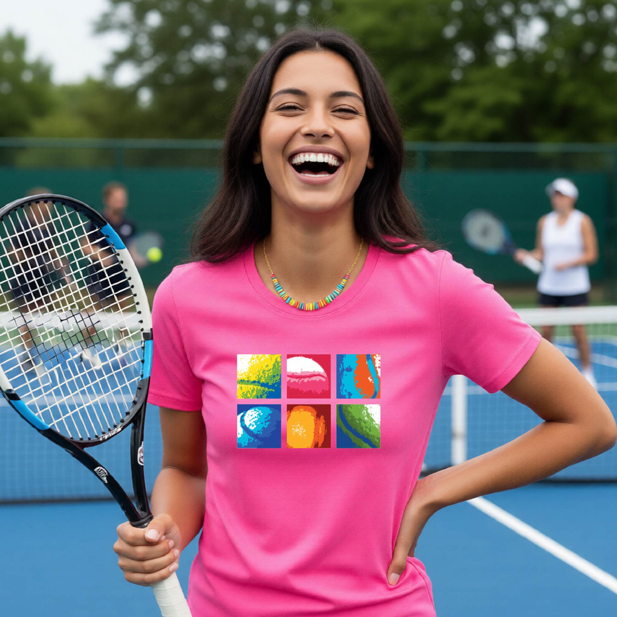 A smiling woman on a blue tennis court holds a tennis racket and wears a pink short-sleeve T-shirt with a colorful 2x3 grid graphic on the chest of tennis balls. Other tennis players are blurred in the background.