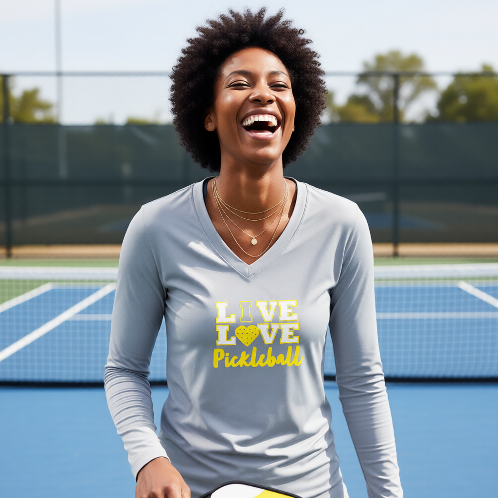 A woman laughing on a pickleball court wearing a light gray long-sleeve V-neck shirt with 'LOVE LOVE Pickleball' printed in yellow on the front with a pickleball motiff heart replacing the O. 