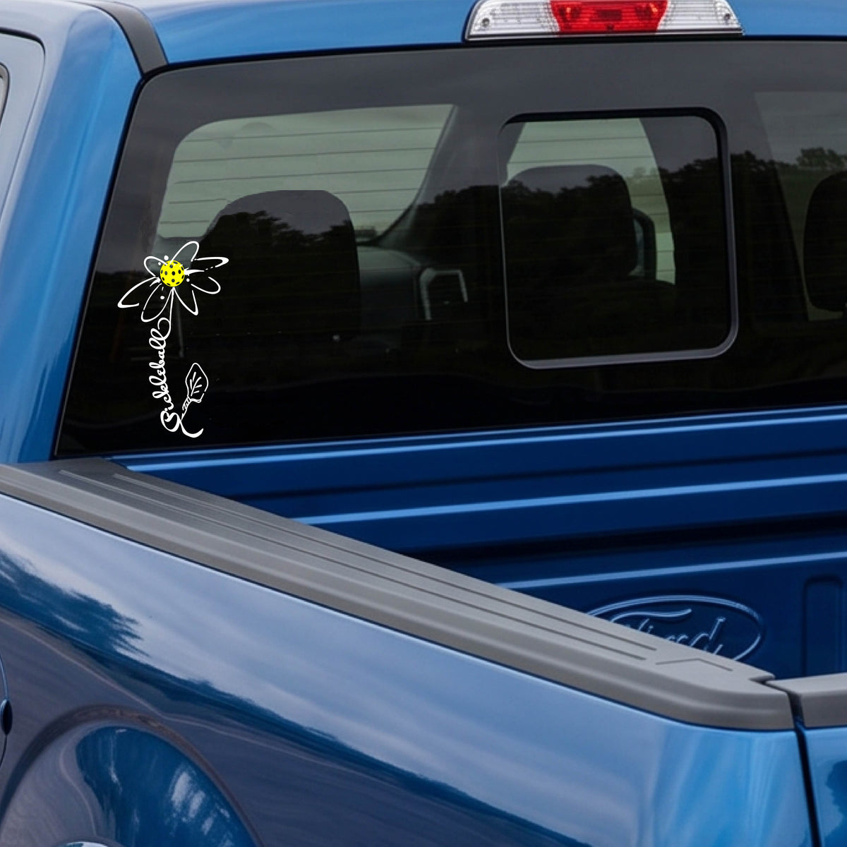 A car window with a white pickleball flower with a stem made out of the text 'PICKLEBALL' and the leaf a pickleblal paddle.applied to the glass.