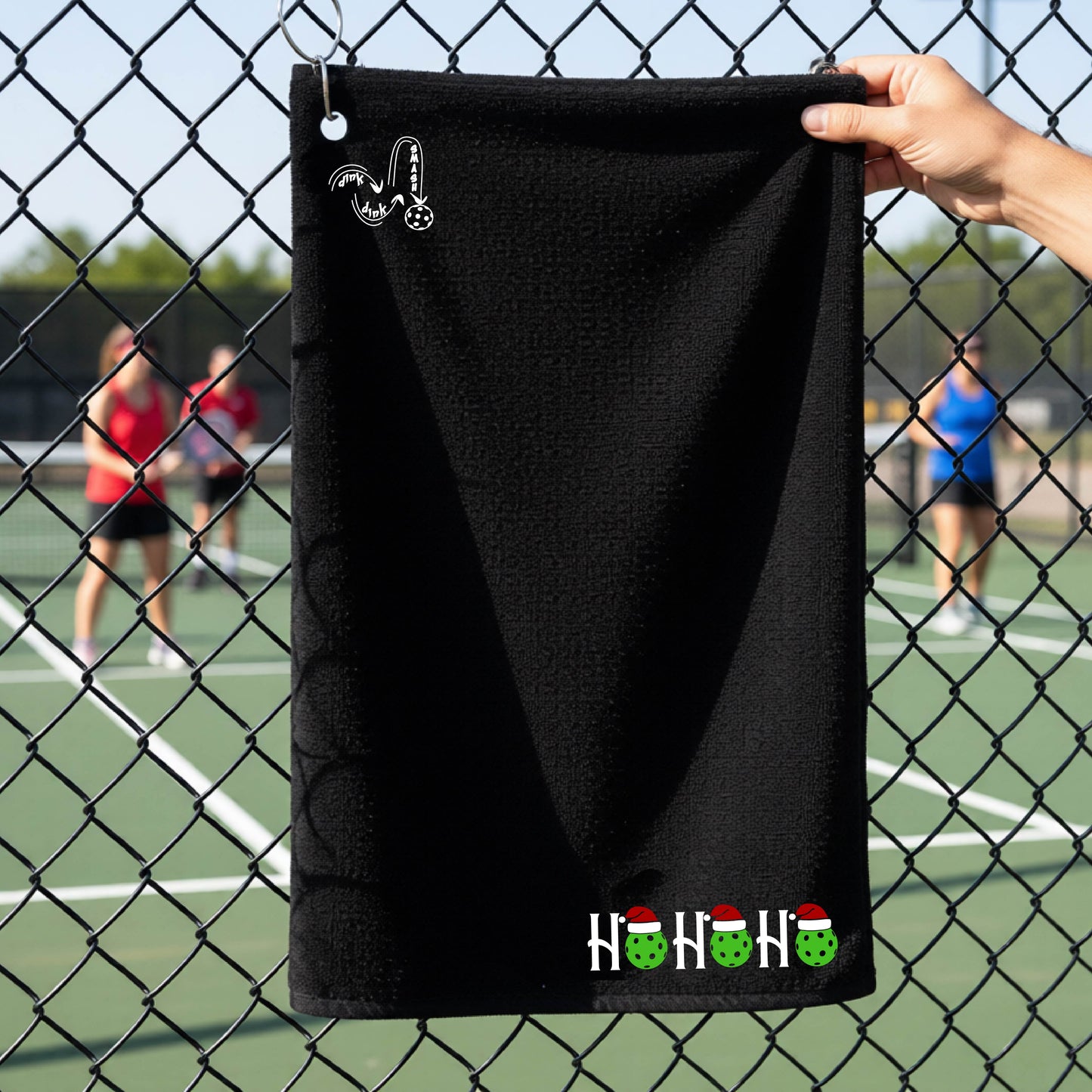 A black towel held up by a hand in front of a chain-link fence at a pickleball court. The top left has a white DINK DINK SMASH logo and a white 'HOHOHO' text design with green pickleballs and red Santa hats near the bottom right.