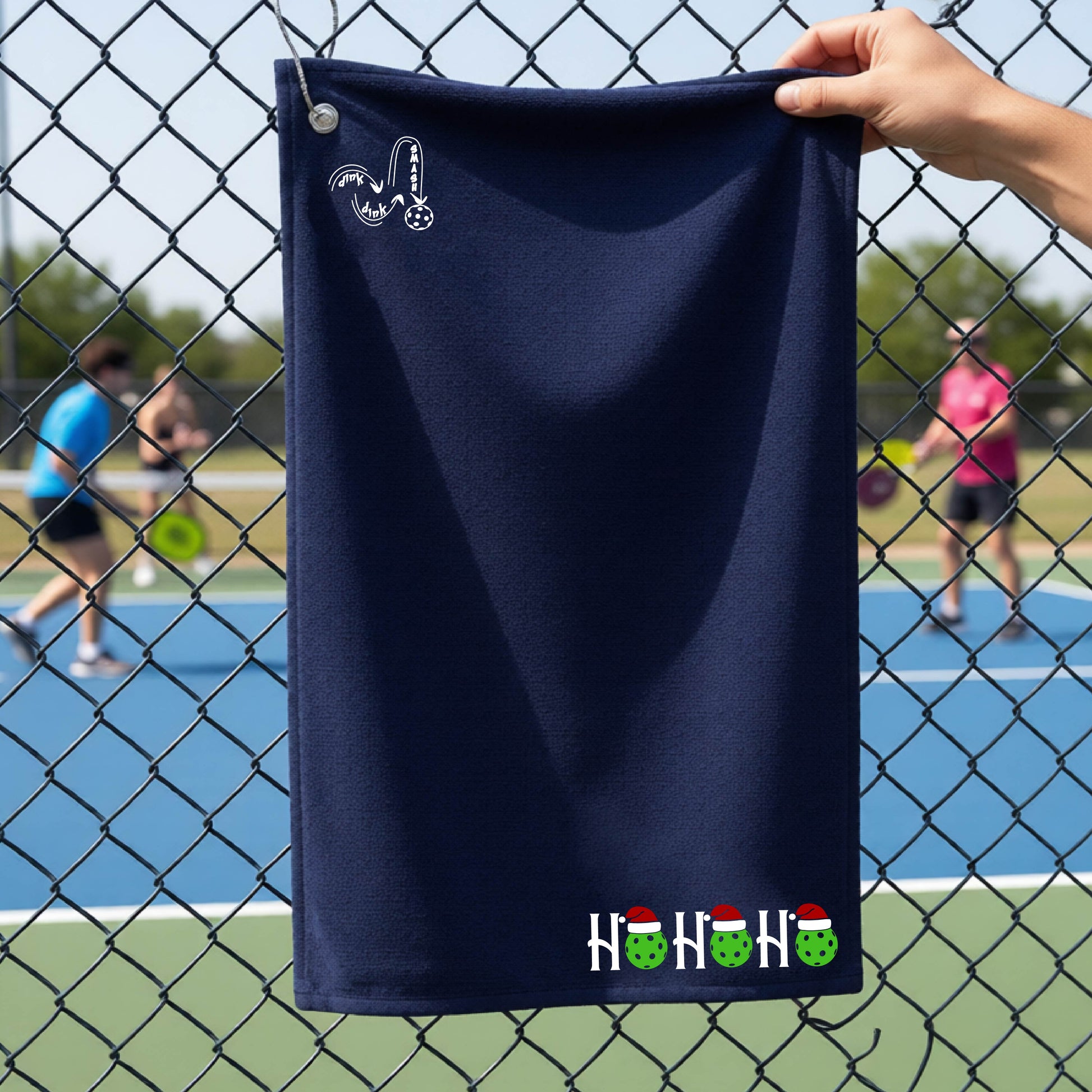 A navy blue towel held up by a hand in front of a chain-link fence at a pickleball court. The top left has a white DINK DINK SMASH logo and a white 'HOHOHO' text design with green pickleballs and red Santa hats near the bottom right.