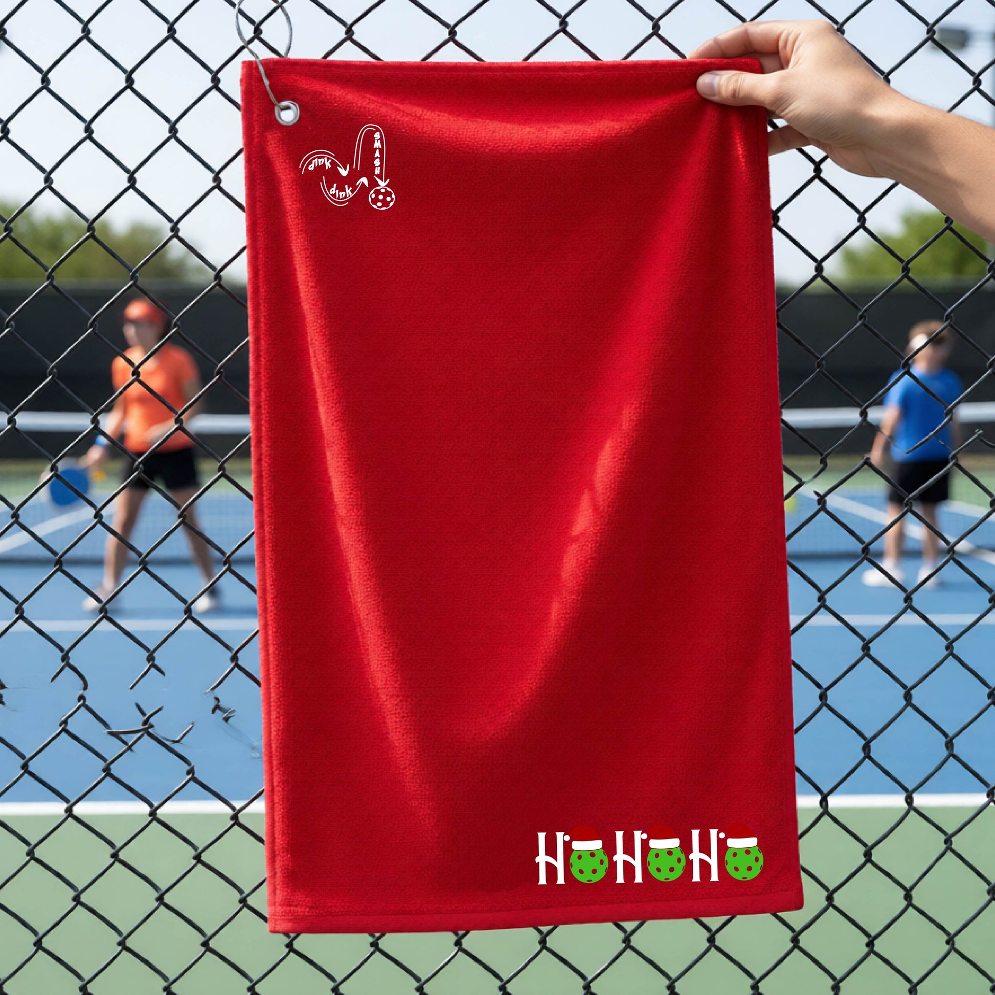 A red towel held up by a hand in front of a chain-link fence at a pickleball court. The top left has a white DINK DINK SMASH logo and a white 'HOHOHO' text design with green pickleballs and red Santa hats near the bottom right.