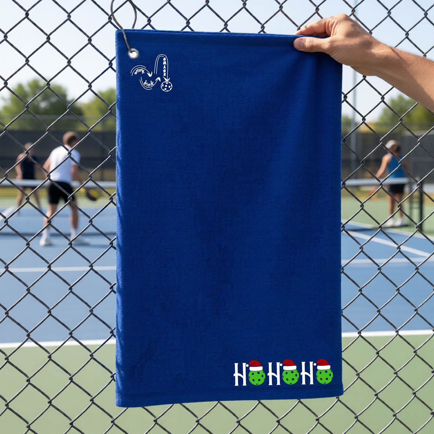 A royal blue towel held up by a hand in front of a chain-link fence at a pickleball court. The top left has a white DINK DINK SMASH logo and a white 'HOHOHO' text design with green pickleballs and red Santa hats near the bottom right.