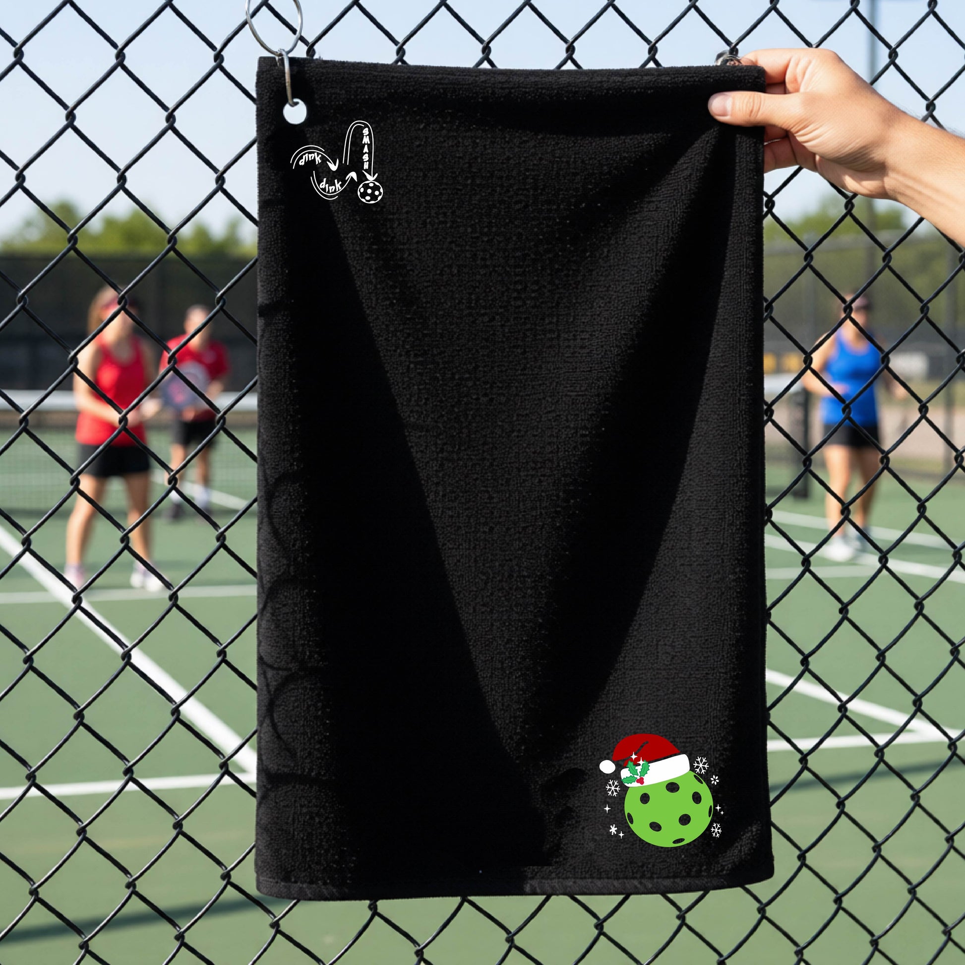 A black towel held up by a hand in front of a chain-link fence at a pickleball court. The top left has a white DINK DINK SMASH logo and in the botttom right corner a green pickleball wearing a santa hat.