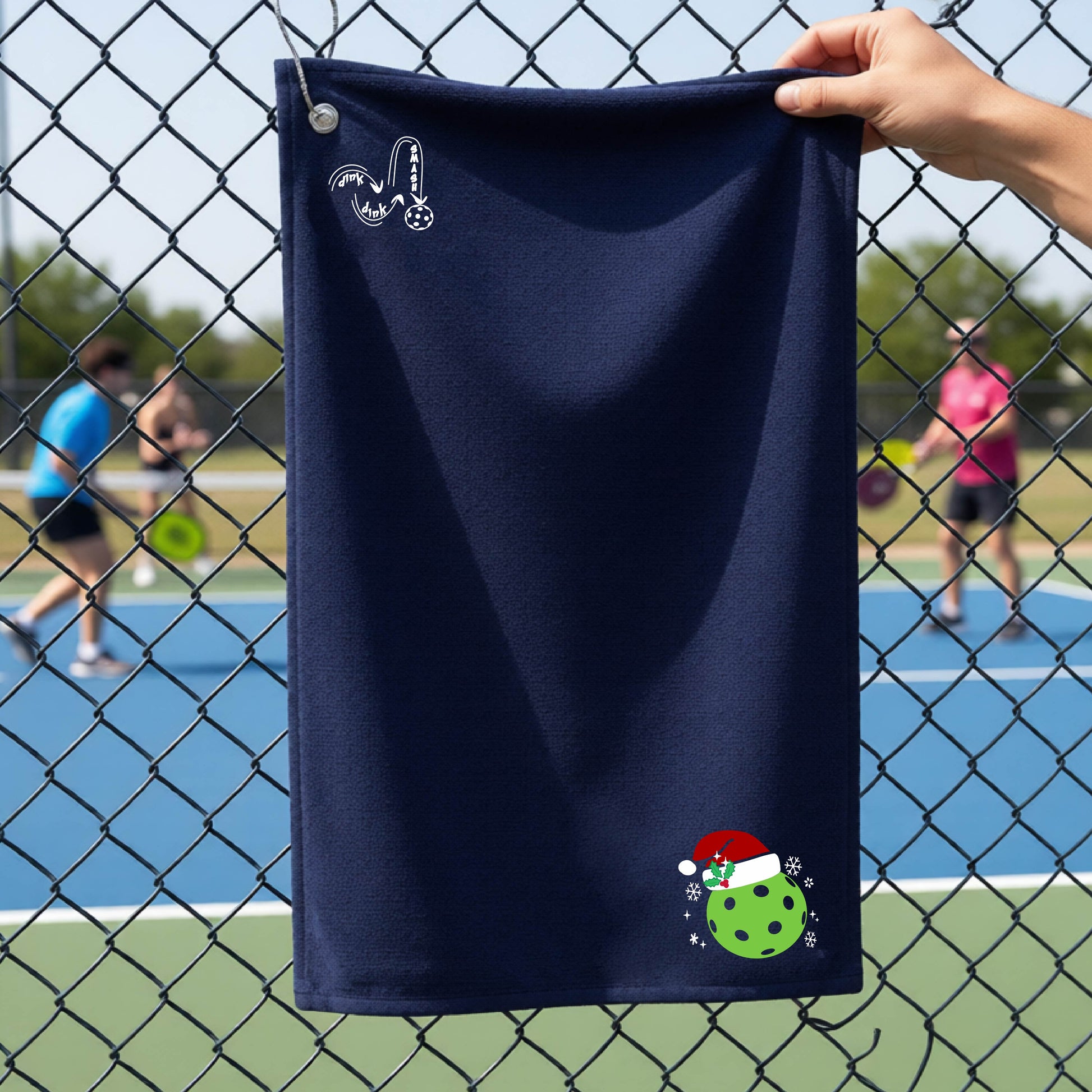 A black towel held up by a hand in front of a chain-link fence at a pickleball court. The top left has a white DINK DINK SMASH logo and in the botttom right corner a green pickleball wearing a santa hat.