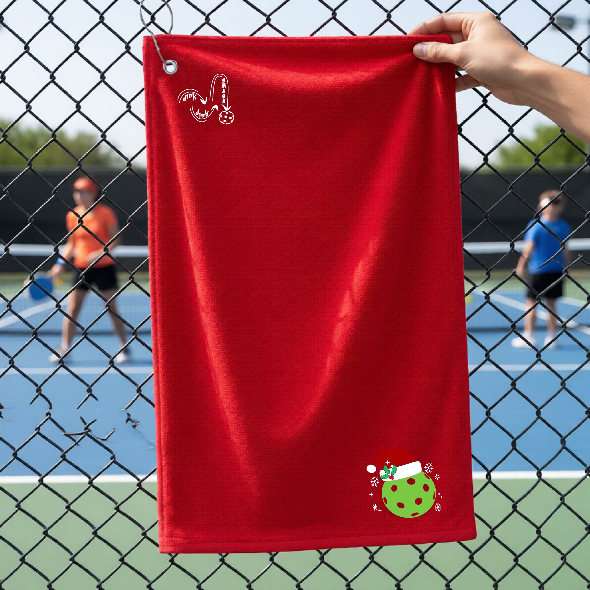 A red towel held up by a hand in front of a chain-link fence at a pickleball court. The top left has a white DINK DINK SMASH logo and in the botttom right corner a green pickleball wearing a santa hat.