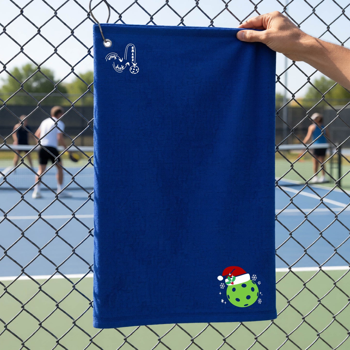 A black towel held up by a hand in front of a chain-link fence at a pickleball court. The top left has a white DINK DINK SMASH logo and in the botttom right corner a green pickleball wearing a santa hat.