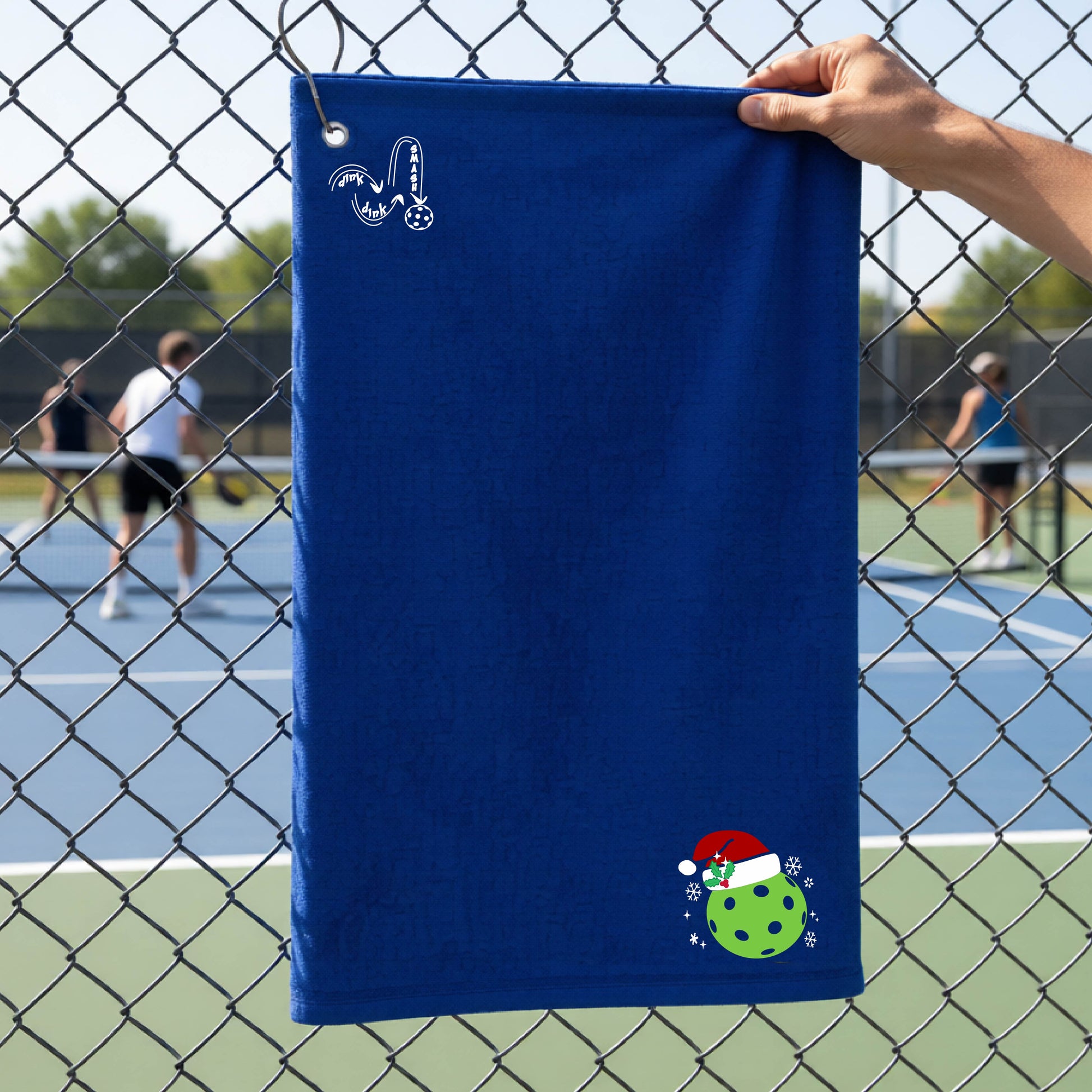 A black towel held up by a hand in front of a chain-link fence at a pickleball court. The top left has a white DINK DINK SMASH logo and in the botttom right corner a green pickleball wearing a santa hat.