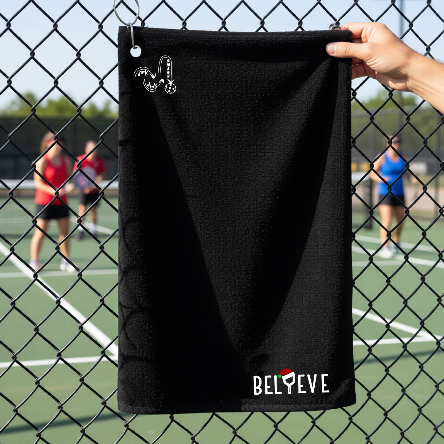 A black towel held up by a hand in front of a chain-link fence at a pickleball court. The top left has a white DINK DINK SMASH logo and in the botttom right corner the text 'BELIEVE' with the I replaced by a pickleball paddle wearing a sant ahat. 
