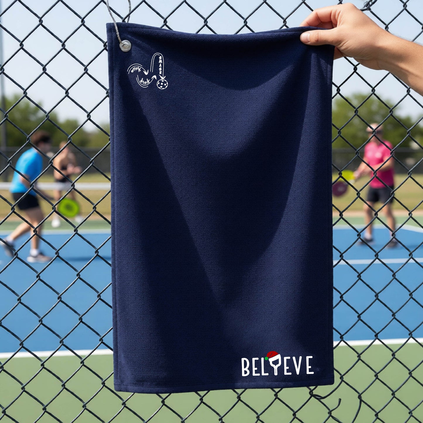 A navy blue towel held up by a hand in front of a chain-link fence at a pickleball court. The top left has a white DINK DINK SMASH logo and in the botttom right corner the text 'BELIEVE' with the I replaced by a pickleball paddle wearing a sant ahat. 