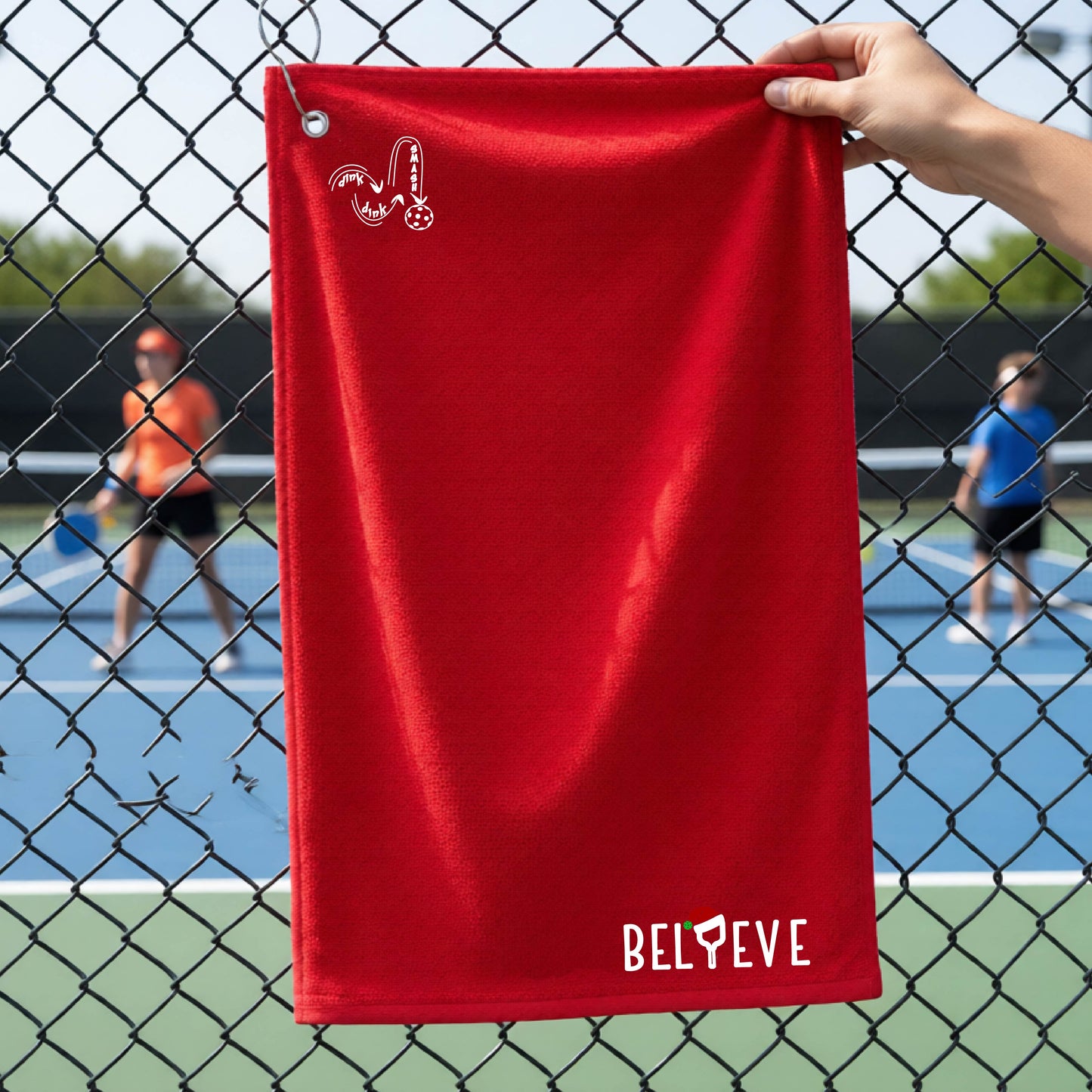 A red towel held up by a hand in front of a chain-link fence at a pickleball court. The top left has a white DINK DINK SMASH logo and in the botttom right corner the text 'BELIEVE' with the I replaced by a pickleball paddle wearing a sant ahat. 