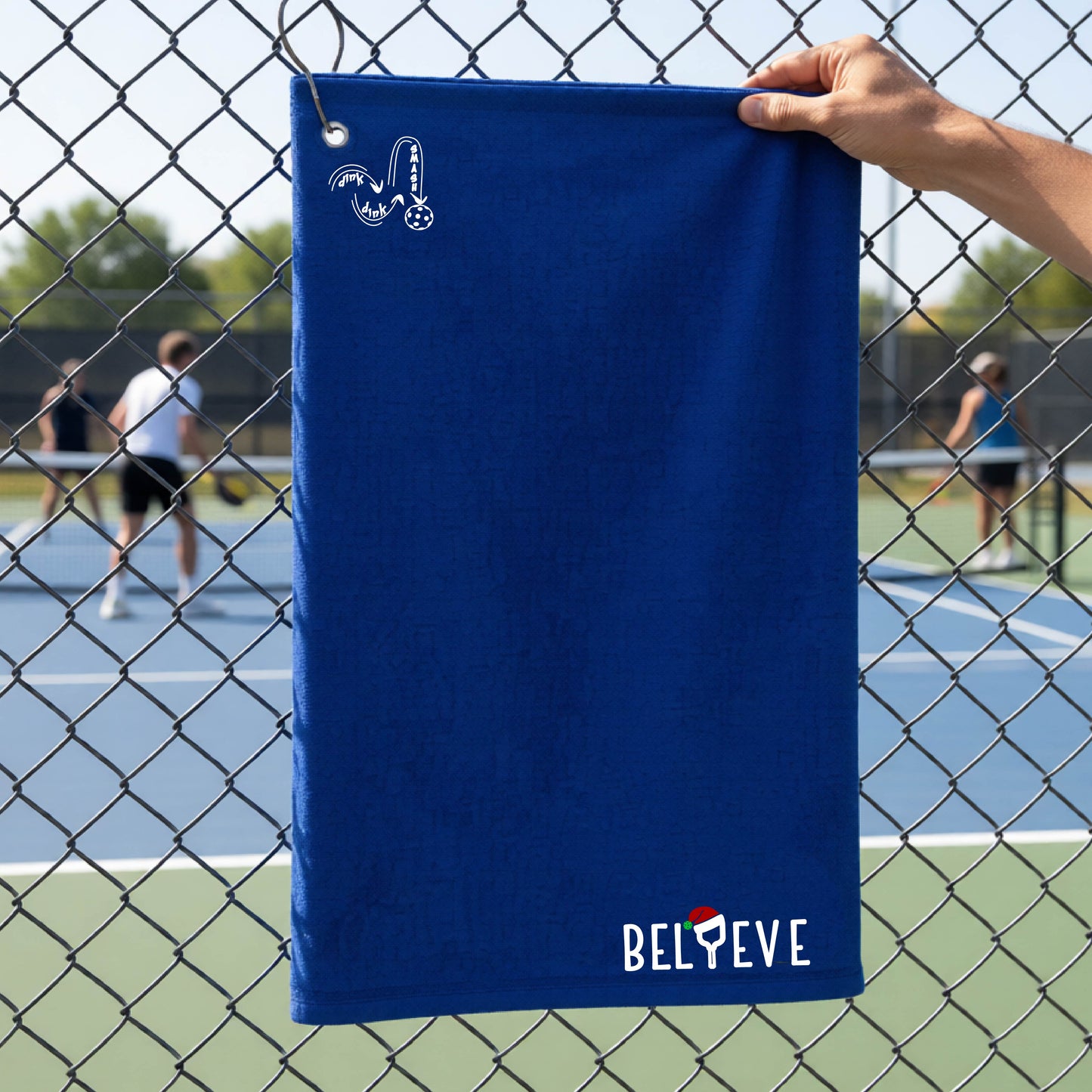 A royal blue towel held up by a hand in front of a chain-link fence at a pickleball court. The top left has a white DINK DINK SMASH logo and in the botttom right corner the text 'BELIEVE' with the I replaced by a pickleball paddle wearing a sant ahat. 