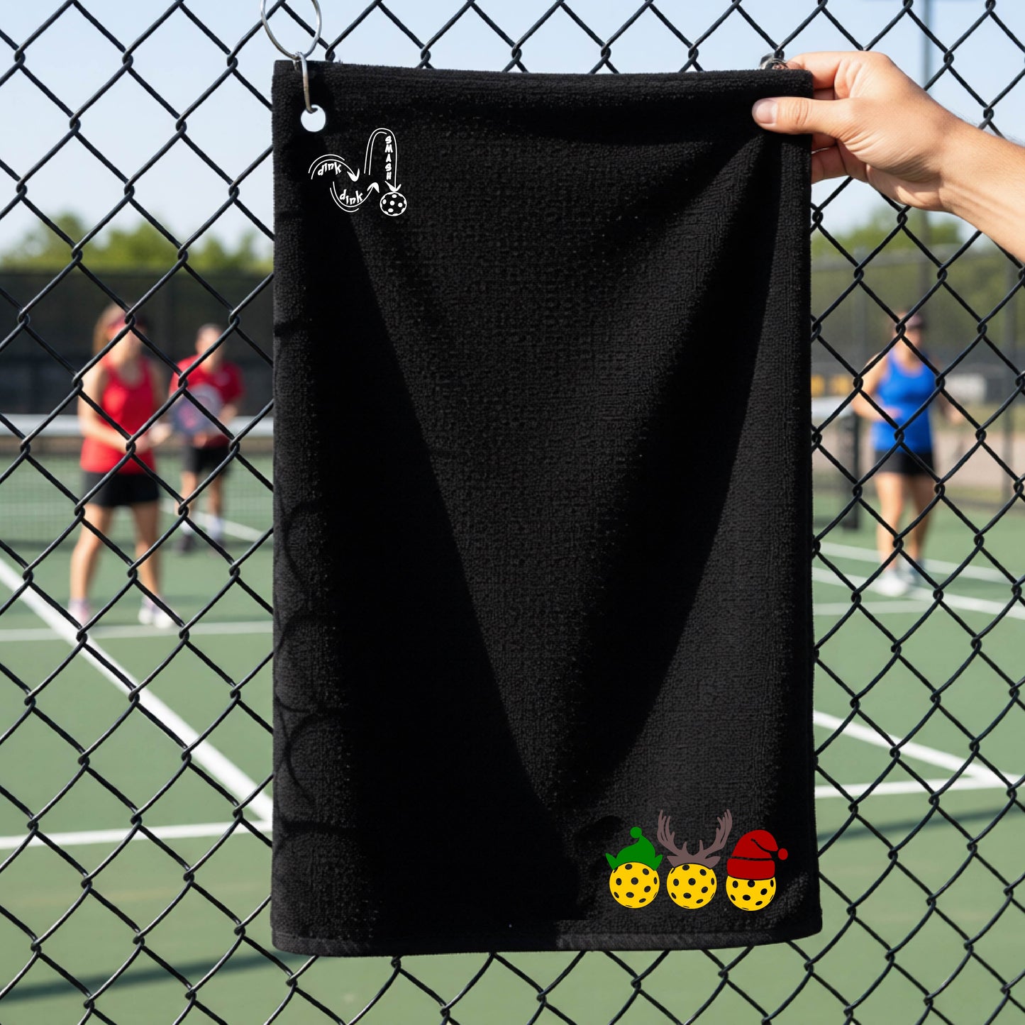 A black towel held up by a hand in front of a chain-link fence at a pickleball court. The top left has a white DINK DINK SMASH logo and a three pickleballs wearing Christmas hats on the bottom right. 