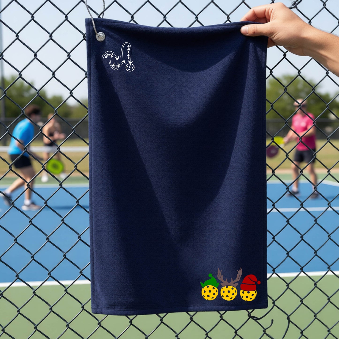 A navy blue towel held up by a hand in front of a chain-link fence at a pickleball court. The top left has a white DINK DINK SMASH logo and a three pickleballs wearing Christmas hats on the bottom right. 