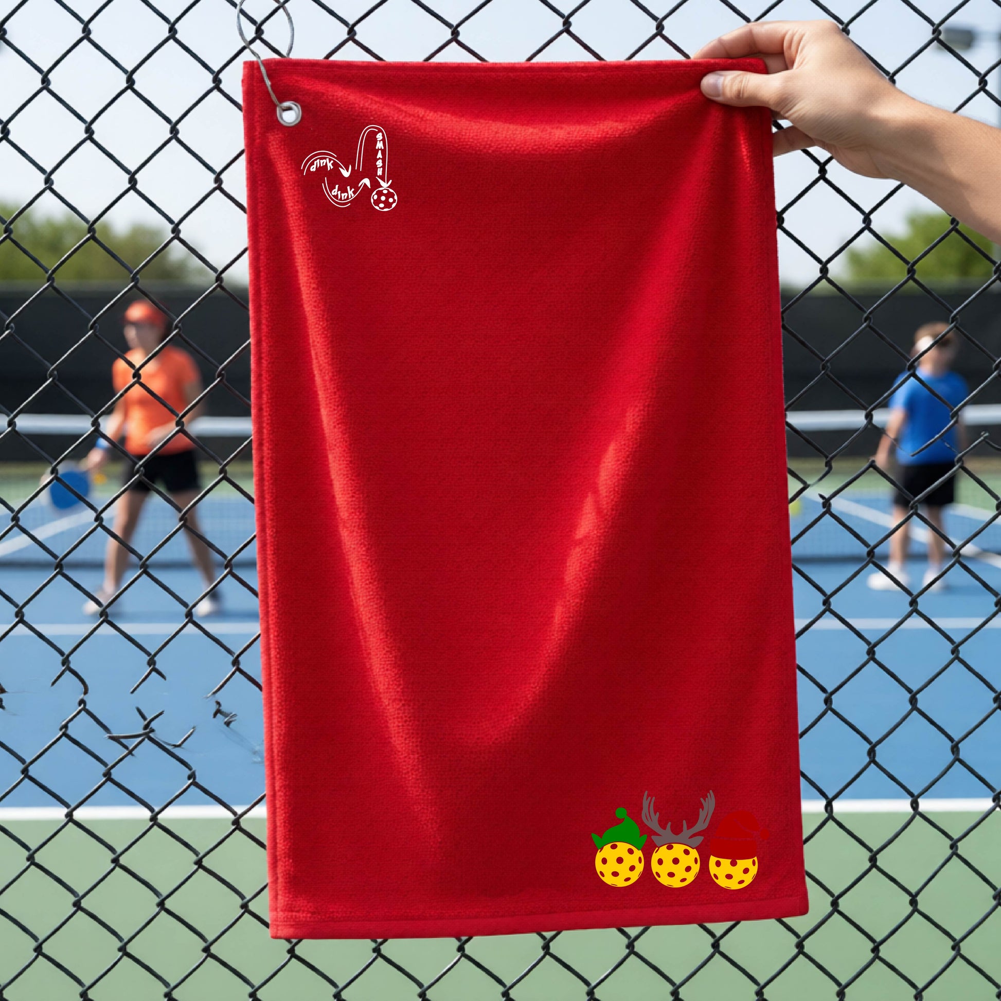 A red towel held up by a hand in front of a chain-link fence at a pickleball court. The top left has a white DINK DINK SMASH logo and a three pickleballs wearing Christmas hats on the bottom right. 