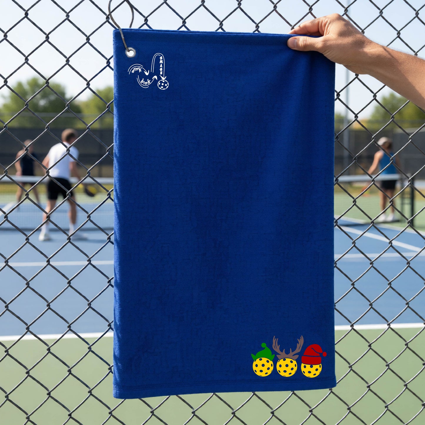 A royal blue towel held up by a hand in front of a chain-link fence at a pickleball court. The top left has a white DINK DINK SMASH logo and a three pickleballs wearing Christmas hats on the bottom right. 