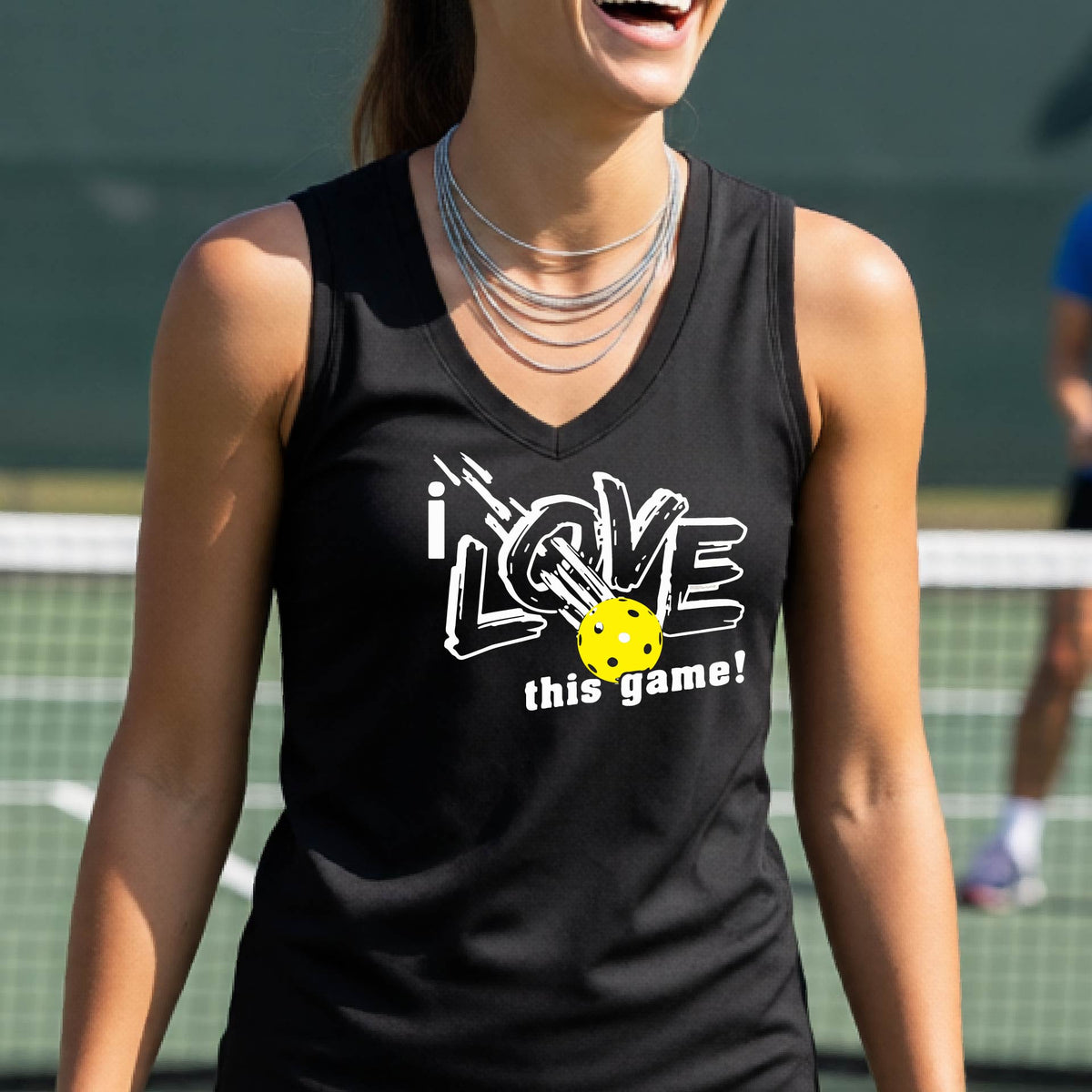 A woman on a pickleball court wearing a black sleeveless tank top with a white graphic that reads 'i LOVE this game!' and a yellow picklaball illustration.