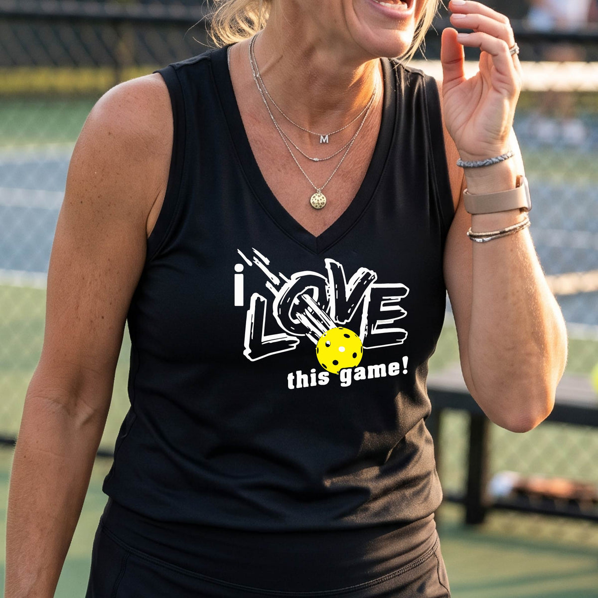 A woman on a pickleball court wearing a black sleeveless tank top with a white graphic that reads 'i LOVE this game!' and a yellow picklaball illustration.