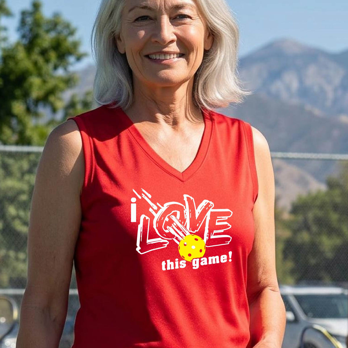 A woman on a pickleball court wearing a red sleeveless tank top with a white graphic that reads 'i LOVE this game!' and a yellow picklaball illustration.