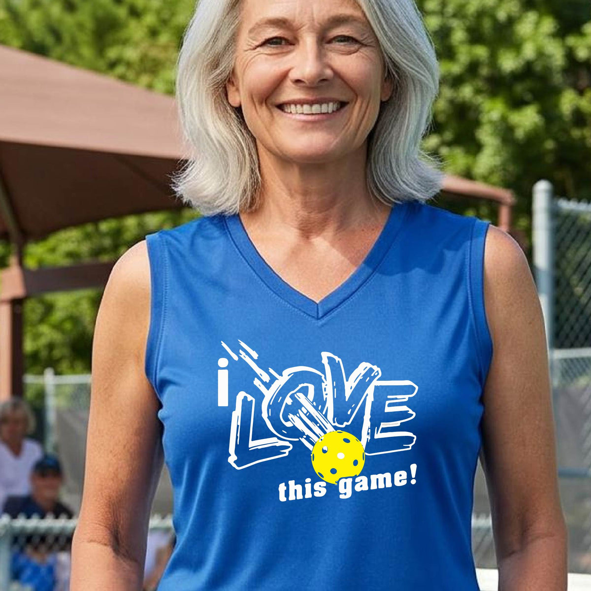 A woman on a pickleball court wearing a royal blue sleeveless tank top with a white graphic that reads 'i LOVE this game!' and a yellow picklaball illustration.
