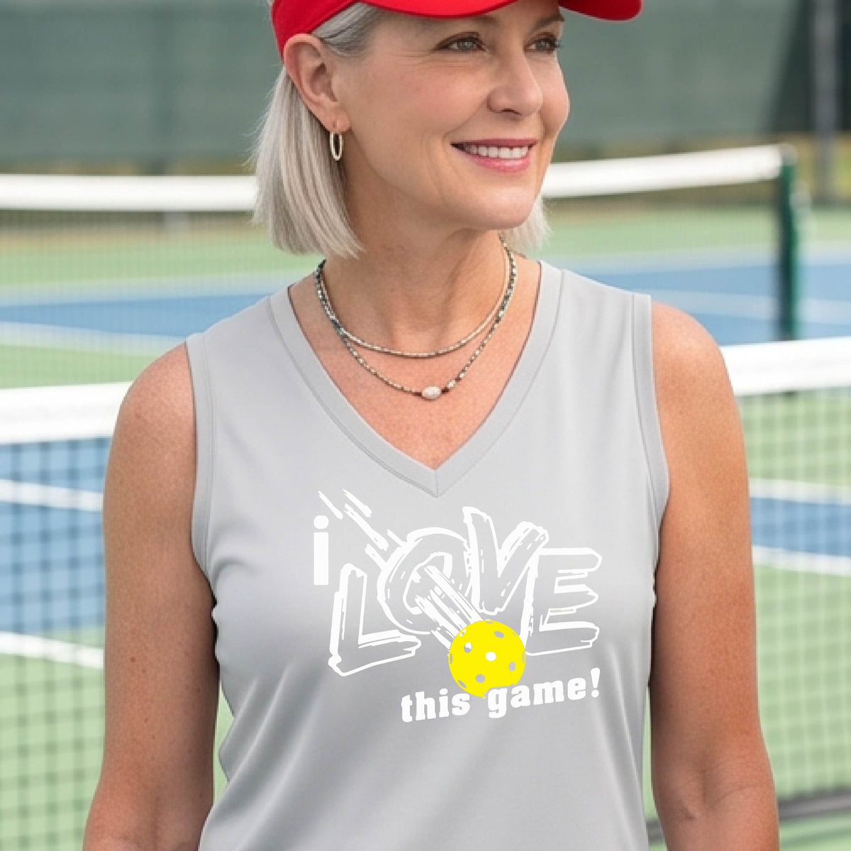 A woman on a pickleball court wearing a silver gray sleeveless tank top with a white graphic that reads 'i LOVE this game!' and a yellow picklaball illustration.