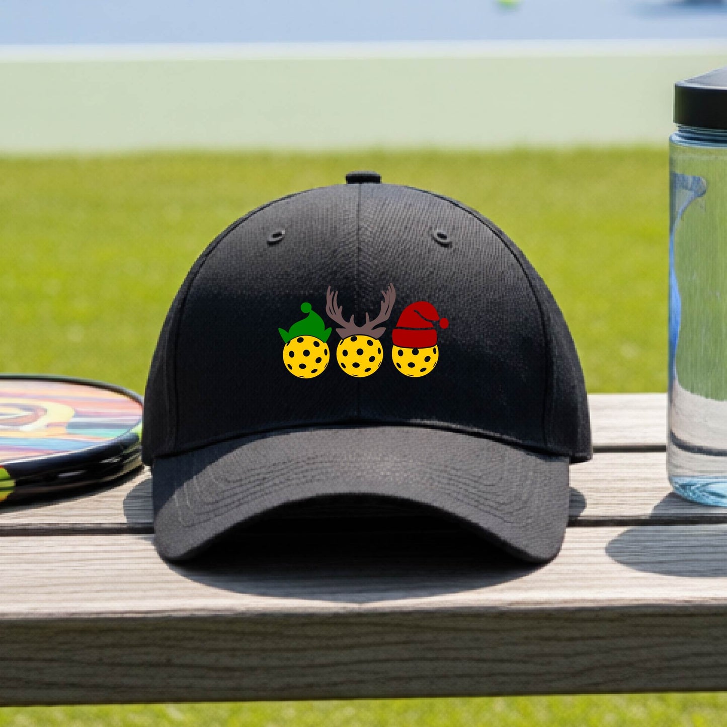 A black baseball hat with a pickleball Christmas graphic of three pickleballs wearing an elf hat, antlers and  santa hat.