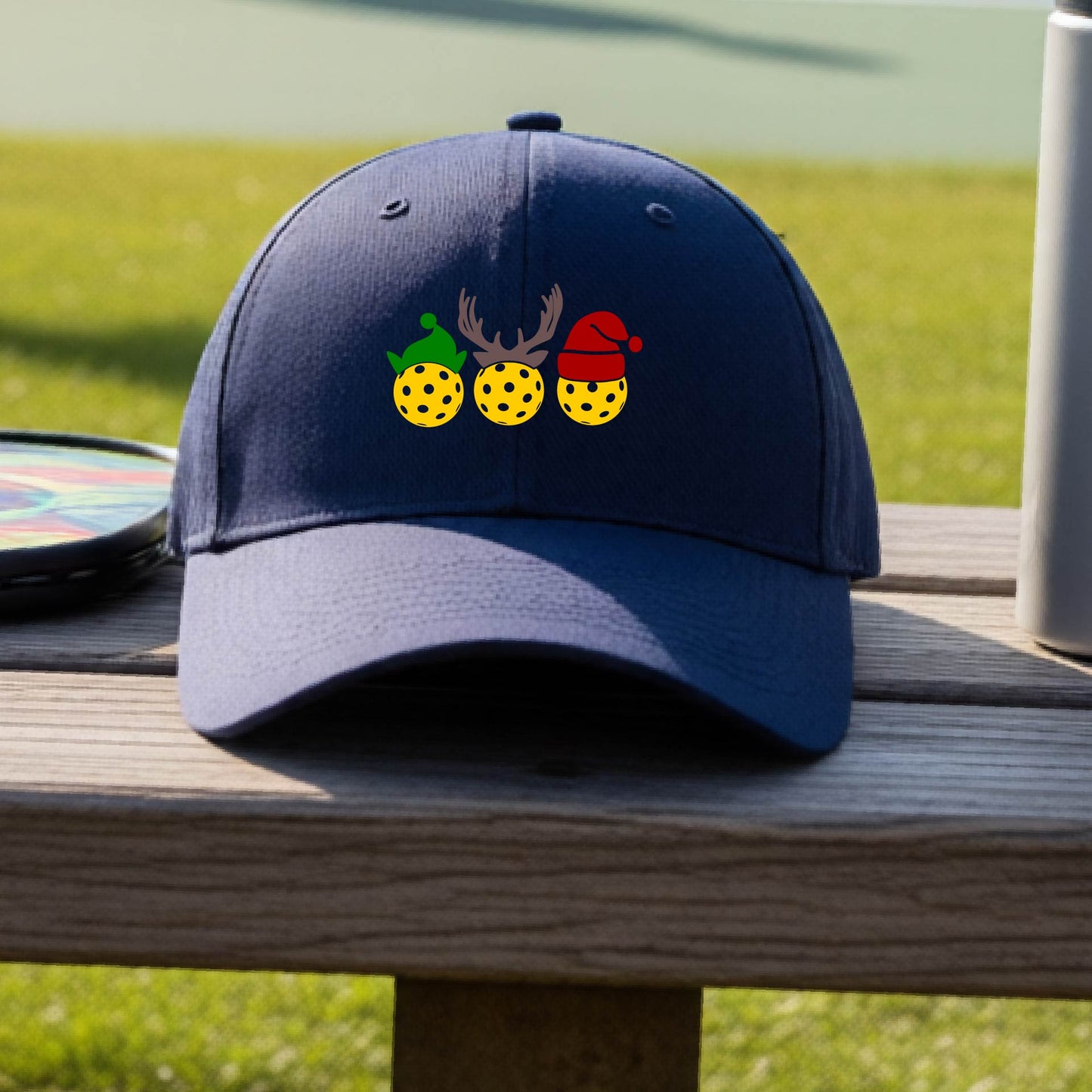 A navy blue baseball hat with a pickleball Christmas graphic of three pickleballs wearing an elf hat, antlers and  santa hat.