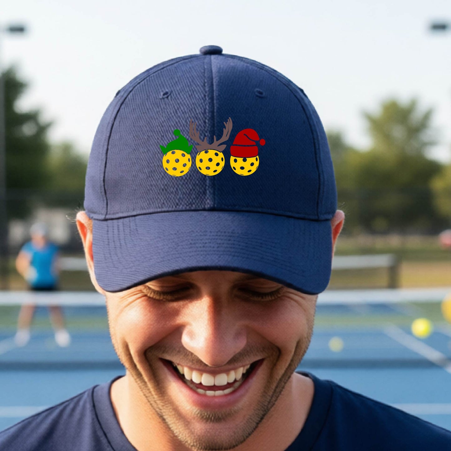 A navy blue baseball hat with a pickleball Christmas graphic of three pickleballs wearing an elf hat, antlers and  santa hat.
