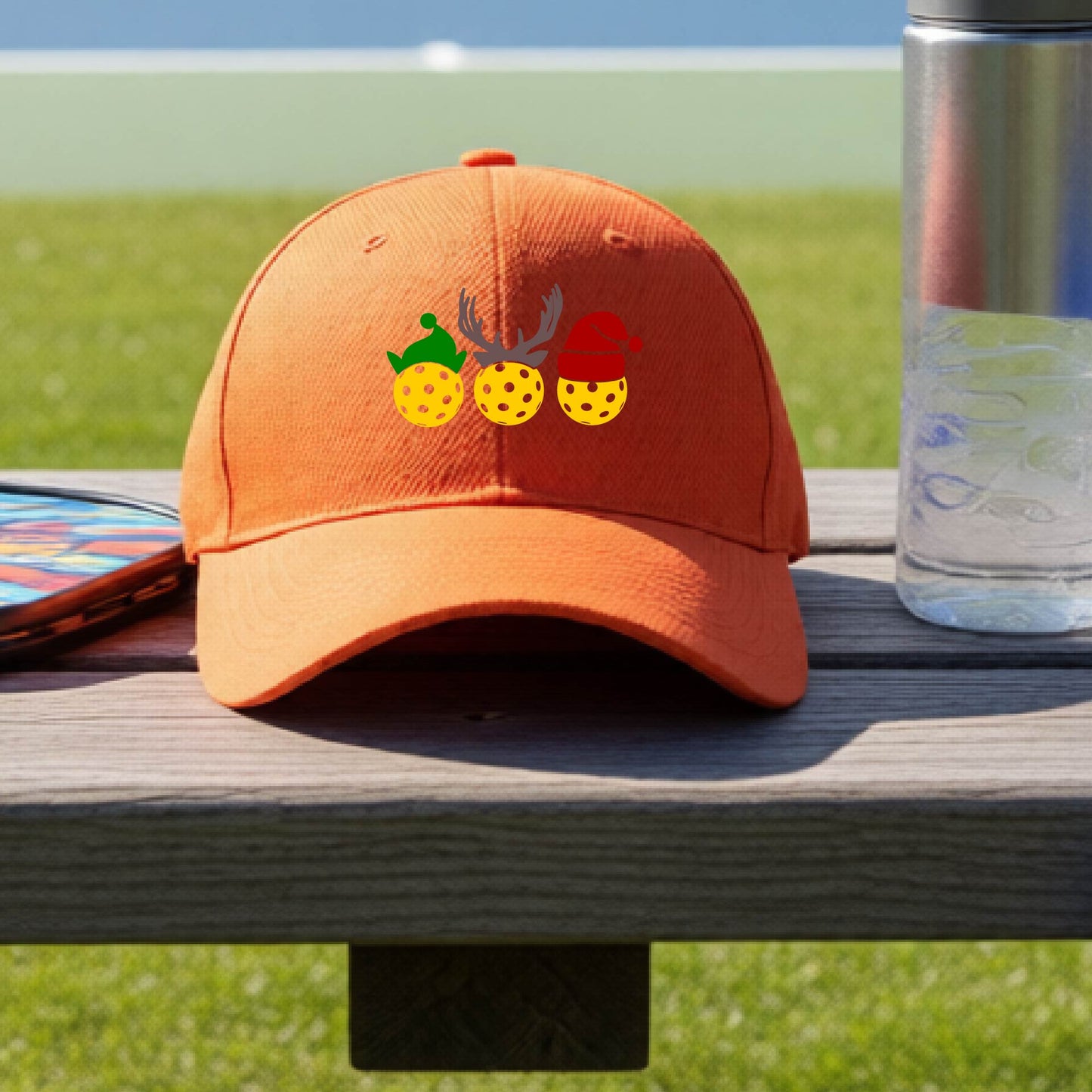 An orangebaseball hat with a pickleball Christmas graphic of three pickleballs wearing an elf hat, antlers and  santa hat.