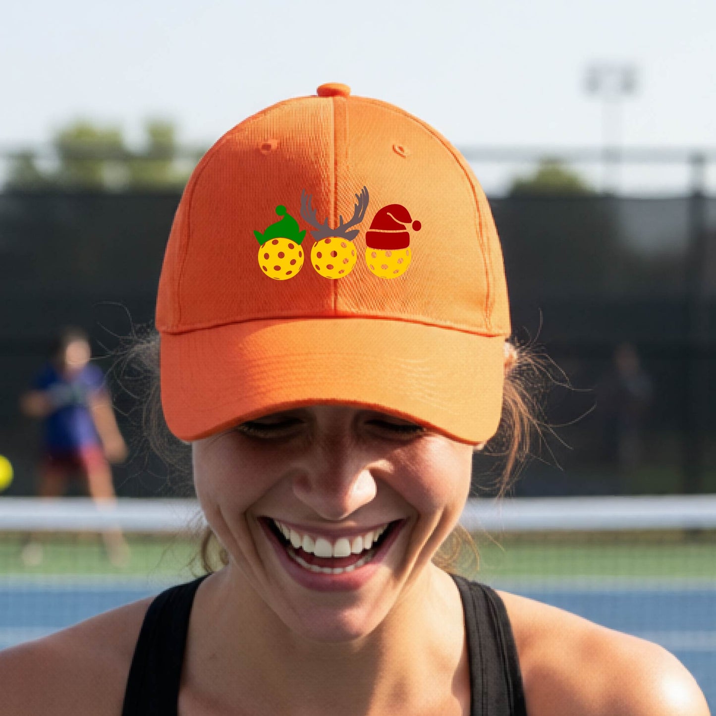 An orange baseball hat with a pickleball Christmas graphic of three pickleballs wearing an elf hat, antlers and  santa hat.