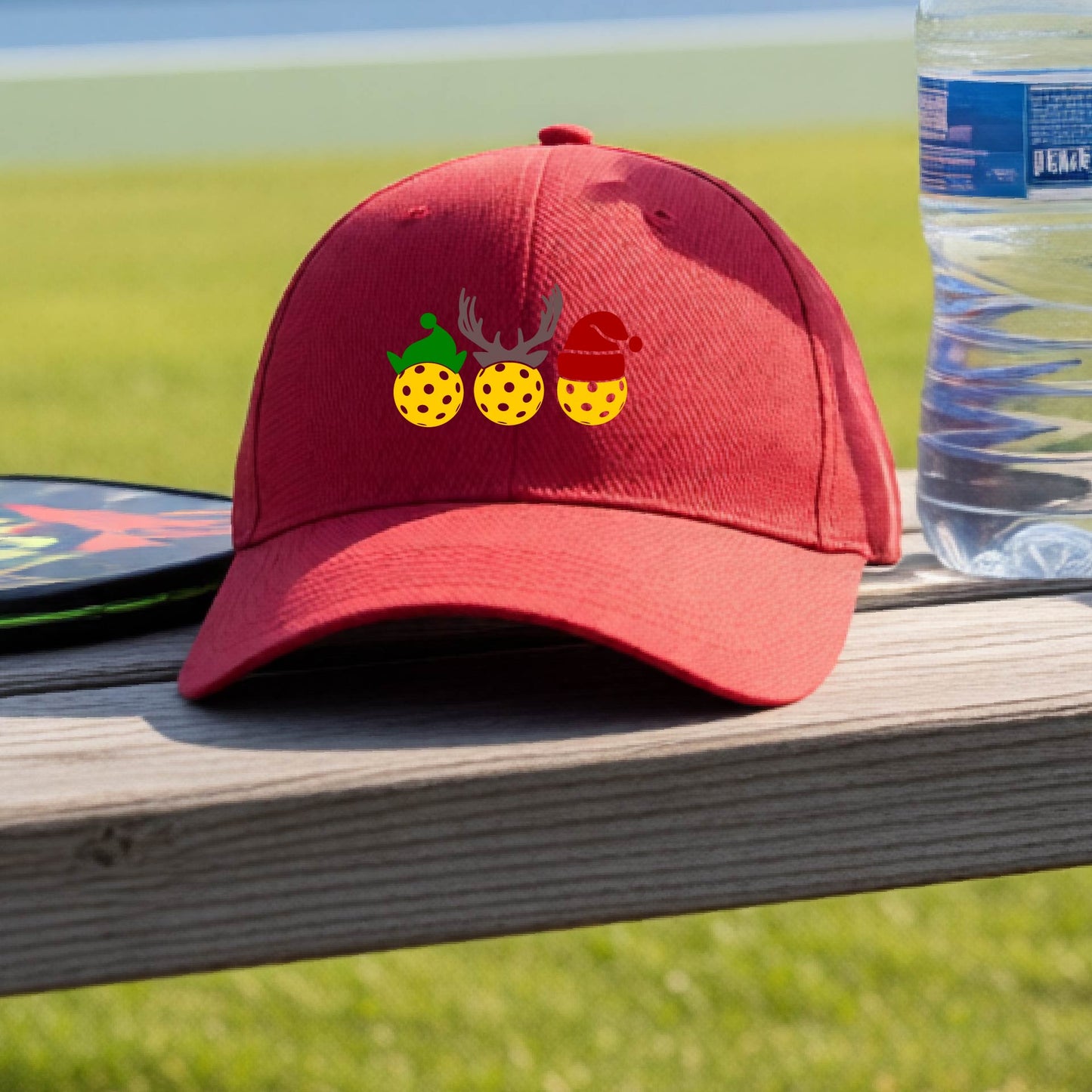 A red baseball hat with a pickleball Christmas graphic of three pickleballs wearing an elf hat, antlers and  santa hat.