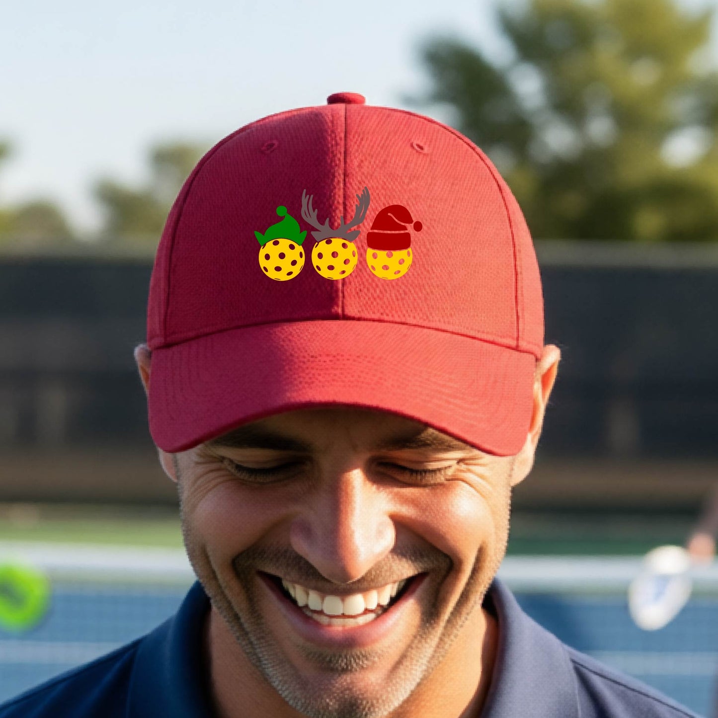 A red baseball hat with a pickleball Christmas graphic of three pickleballs wearing an elf hat, antlers and  santa hat.