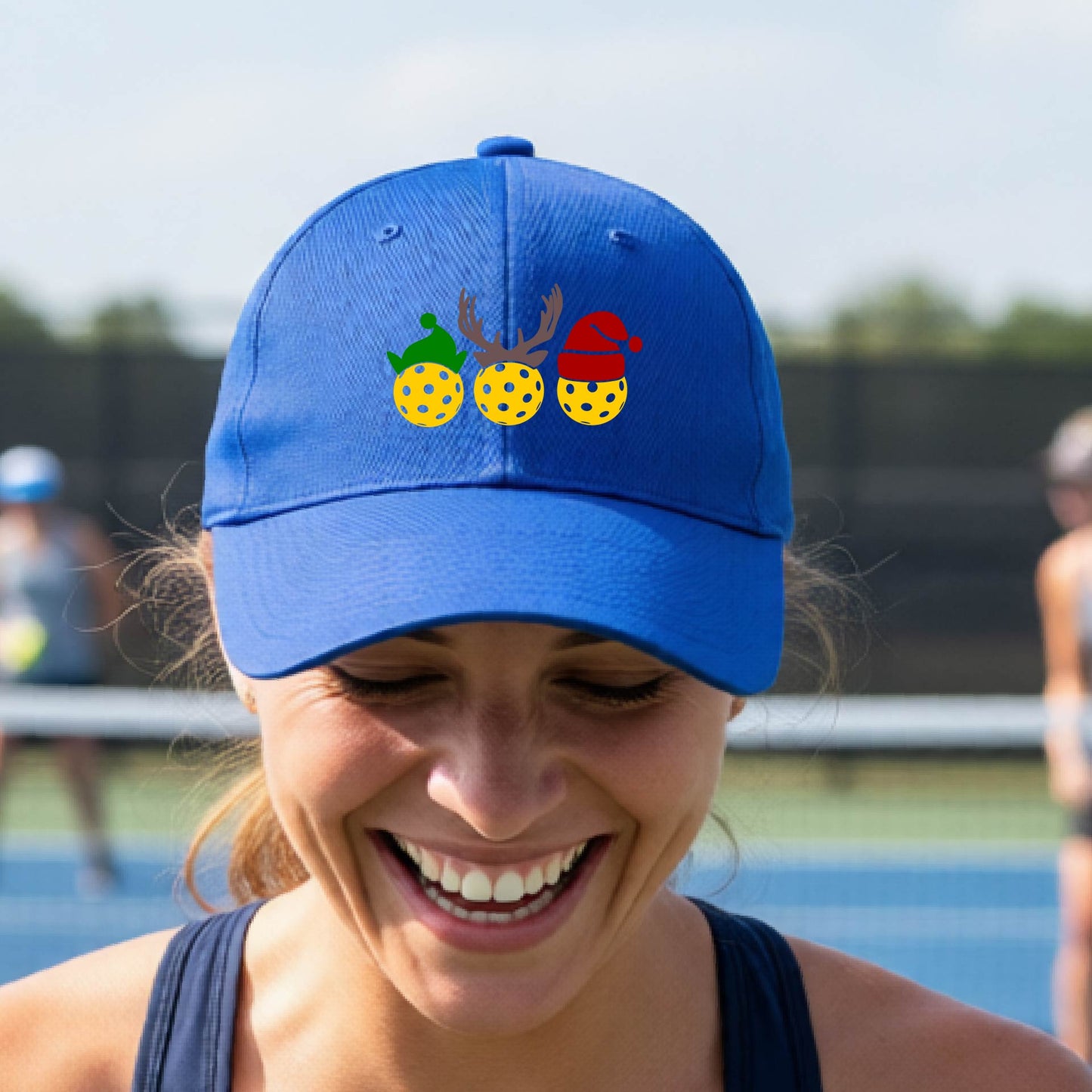 A royal blue baseball hat with a pickleball Christmas graphic of three pickleballs wearing an elf hat, antlers and  santa hat.