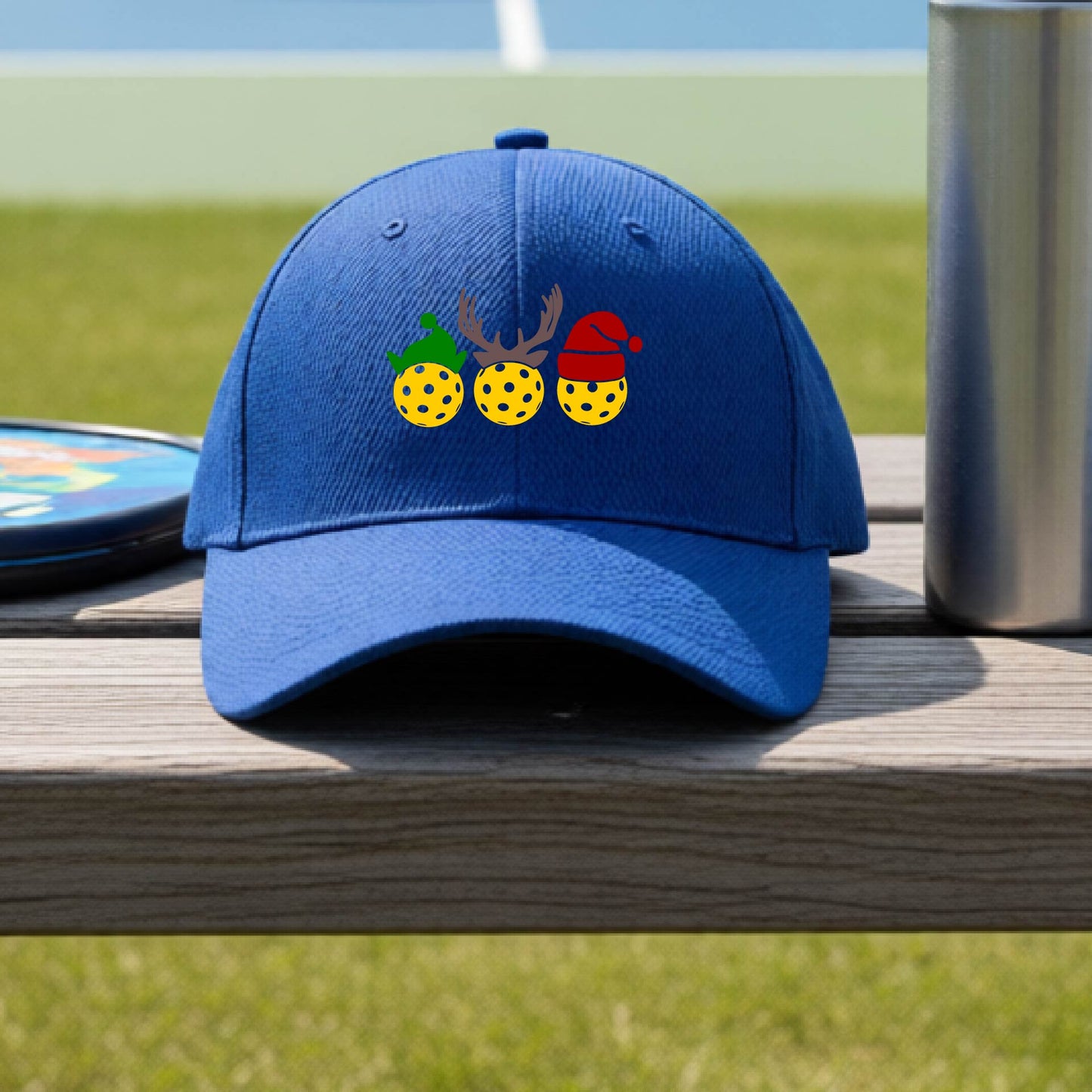 A royal blue baseball hat with a pickleball Christmas graphic of three pickleballs wearing an elf hat, antlers and  santa hat.