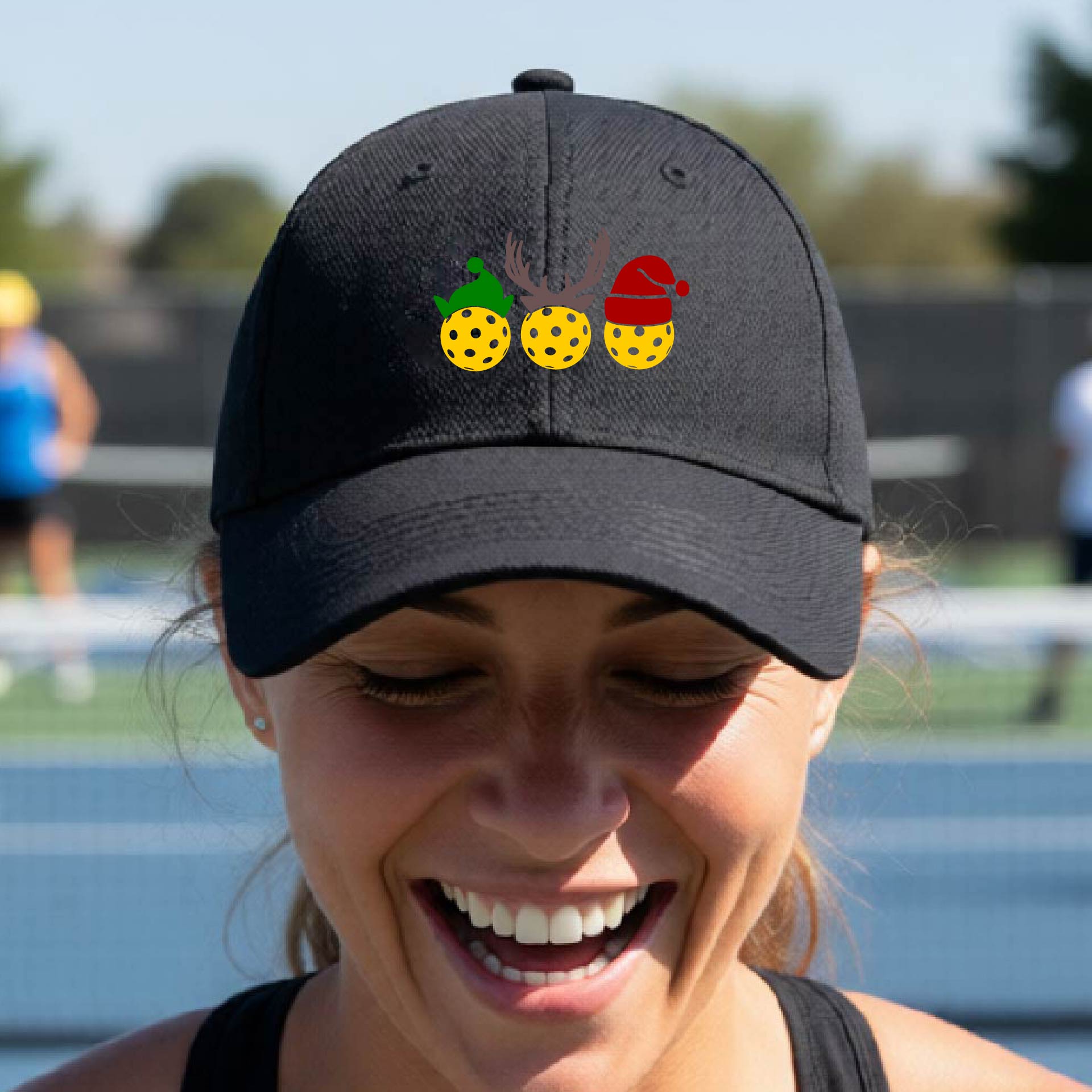A black baseball hat with a pickleball Christmas graphic of three pickleballs wearing an elf hat, antlers and  santa hat.