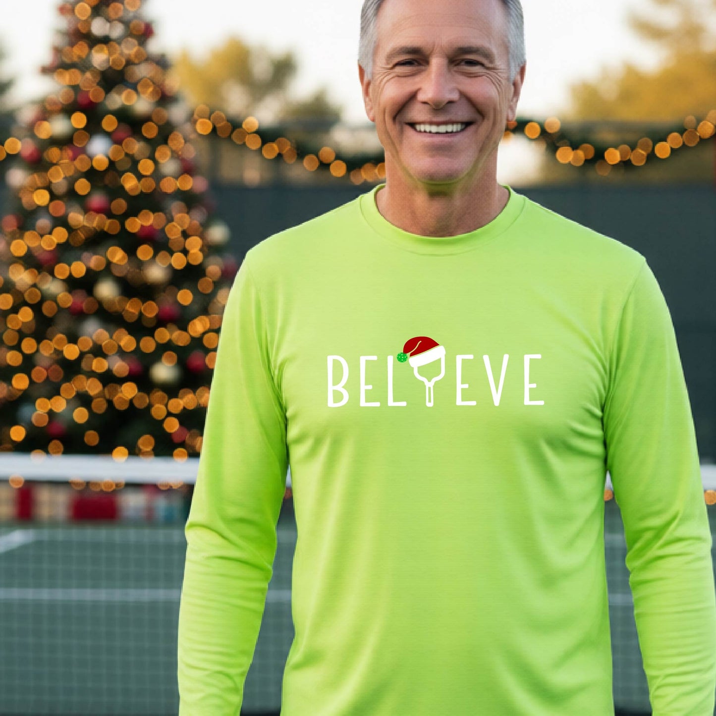 A smiling man wearing a lime green long-sleeve shirt with the word 'BELIEVE' with the I repalced with a pickleball paddle wearing a Santa hat graphic stands on a pickleball court with a Christmas tree and lights n the background.