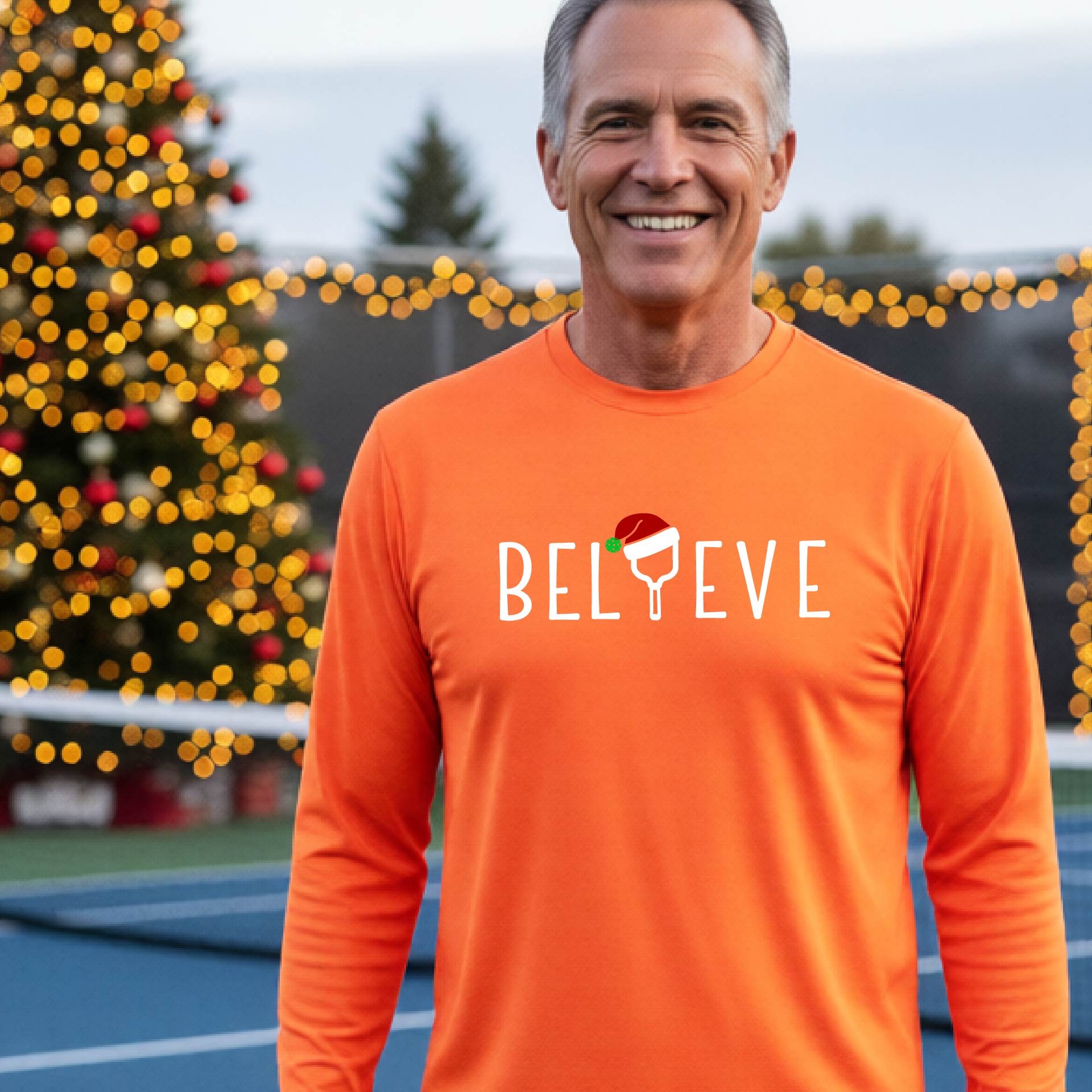 A smiling man wearing a bright orange long-sleeve shirt with the word 'BELIEVE' with the I repalced with a pickleball paddle wearing a Santa hat graphic stands on a pickleball court with a Christmas tree and lights n the background.