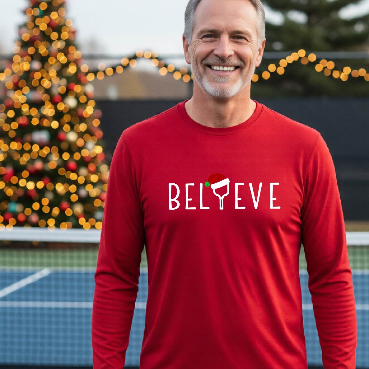 A smiling man wearing a red long-sleeve shirt with the word 'BELIEVE' with the I repalced with a pickleball paddle wearing a Santa hat graphic stands on a pickleball court with a Christmas tree and lights n the background.
