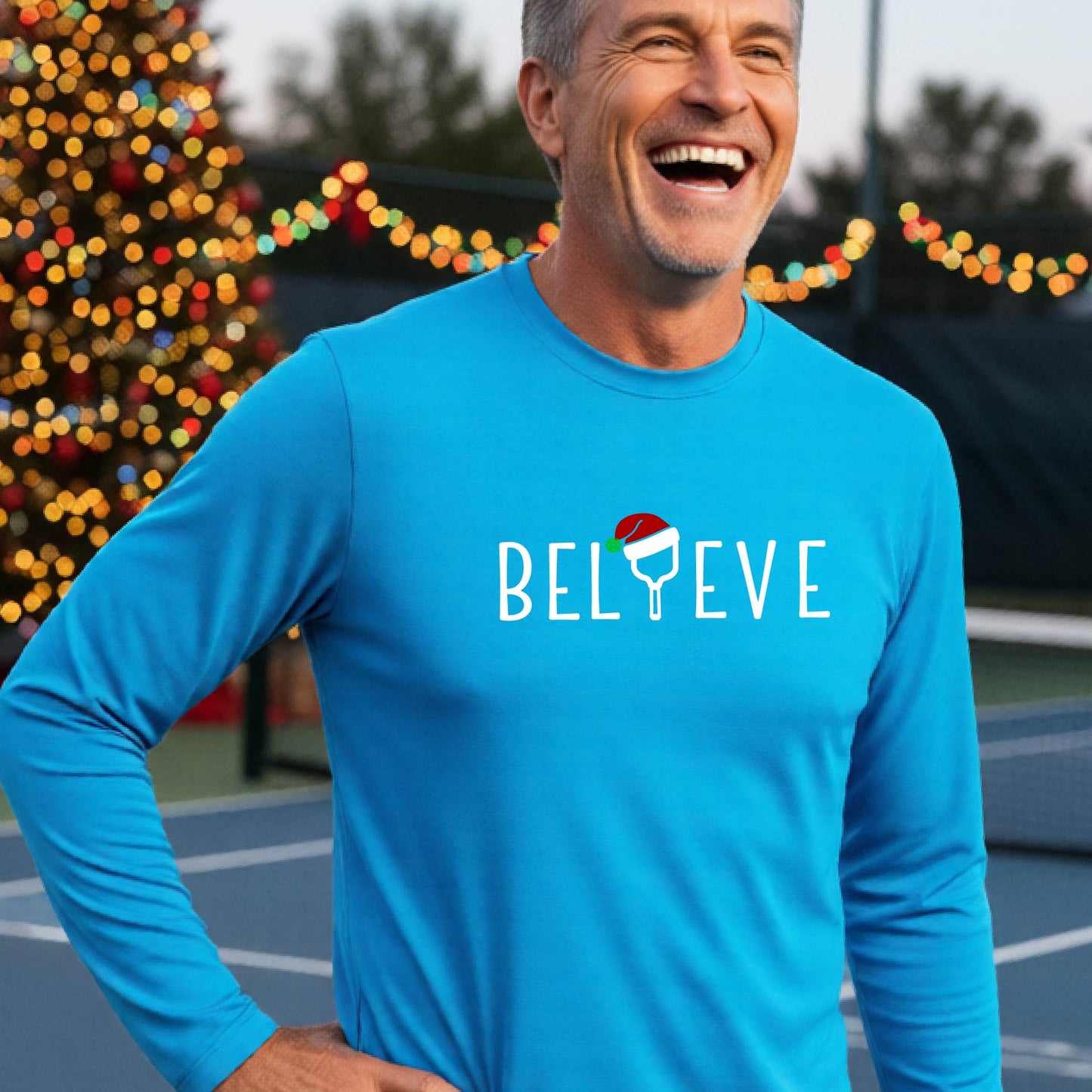 A smiling man wearing a bright blue long-sleeve shirt with the word 'BELIEVE' with the I repalced with a pickleball paddle wearing a Santa hat graphic stands on a pickleball court with a Christmas tree and lights n the background.
