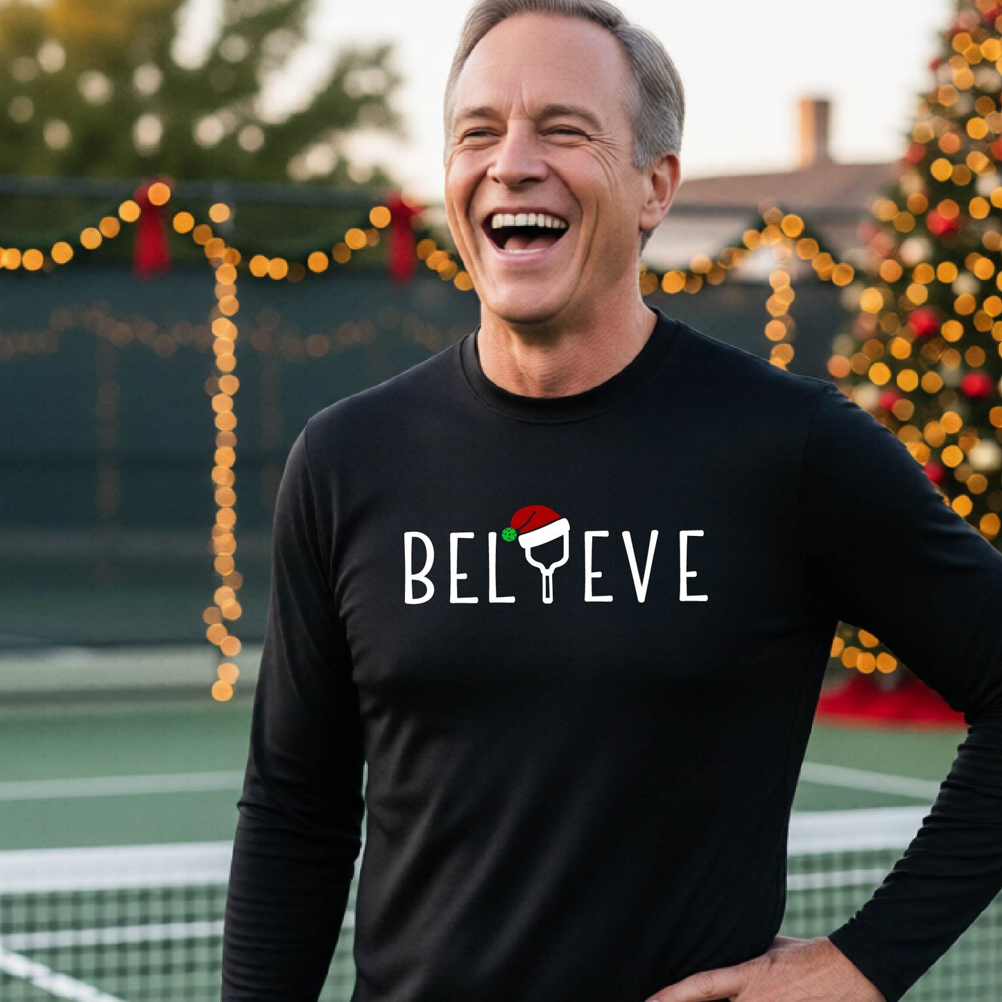 A smiling man wearing a black long-sleeve shirt with the word 'BELIEVE' with the I repalced with a pickleball paddle wearing a Santa hat graphic stands on a pickleball court with a Christmas tree and lights n the background.