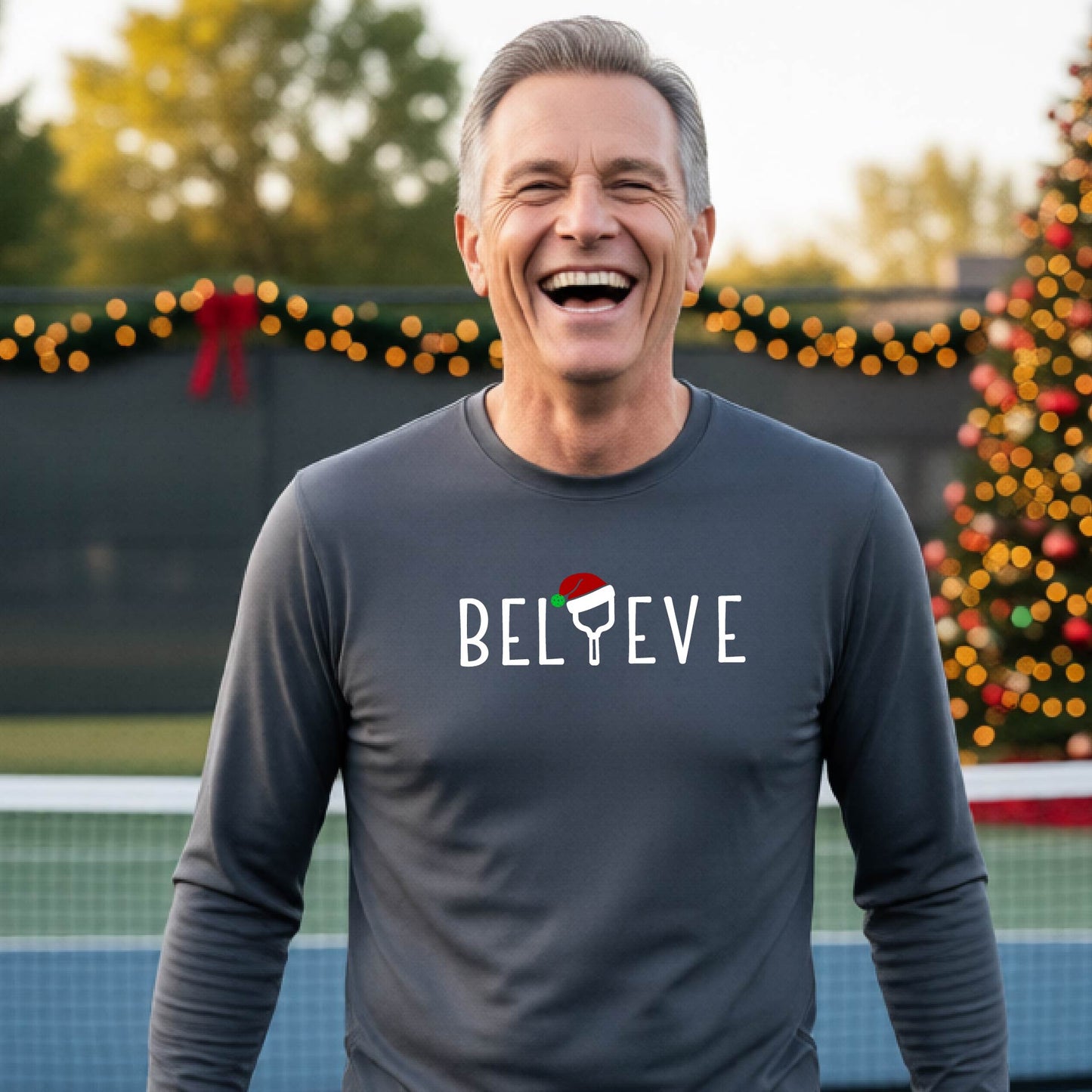 A smiling man wearing a gray long-sleeve shirt with the word 'BELIEVE' with the I repalced with a pickleball paddle wearing a Santa hat graphic stands on a pickleball court with a Christmas tree and lights n the background.
