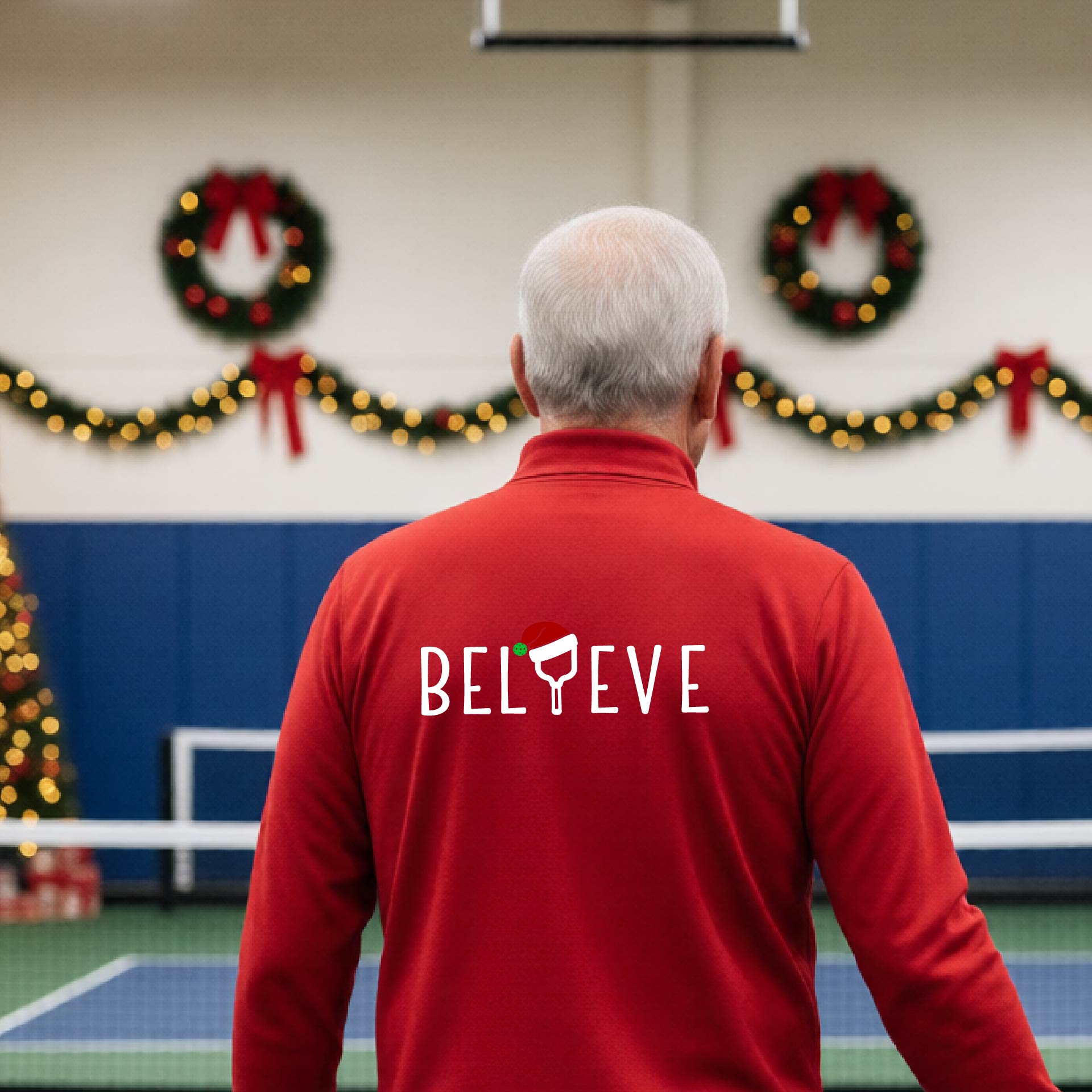 A smiling man wearing a red quarter zip long-sleeveshirt with the word 'BELIEVE' with the I repalced with a pickleball paddle wearing a Santa hat graphic stands on a pickleball court with a Christmas tree iand lights n the background.