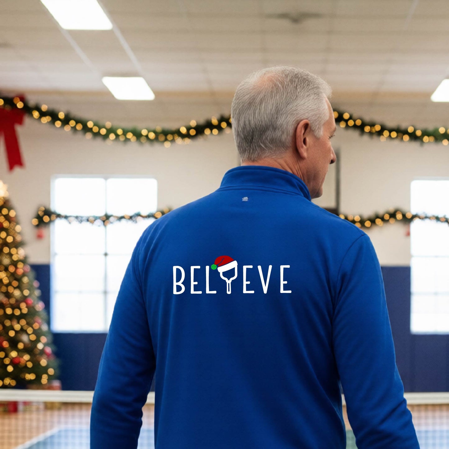 A smiling man wearing a royal blue quarter zip long-sleeveshirt with the word 'BELIEVE' with the I repalced with a pickleball paddle wearing a Santa hat graphic stands on a pickleball court with a Christmas tree iand lights n the background.