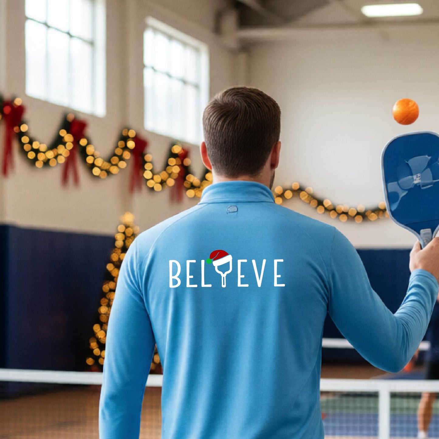 A smiling man wearing a light blue quarter zip long-sleeveshirt with the word 'BELIEVE' with the I repalced with a pickleball paddle wearing a Santa hat graphic stands on a pickleball court with a Christmas tree iand lights n the background.