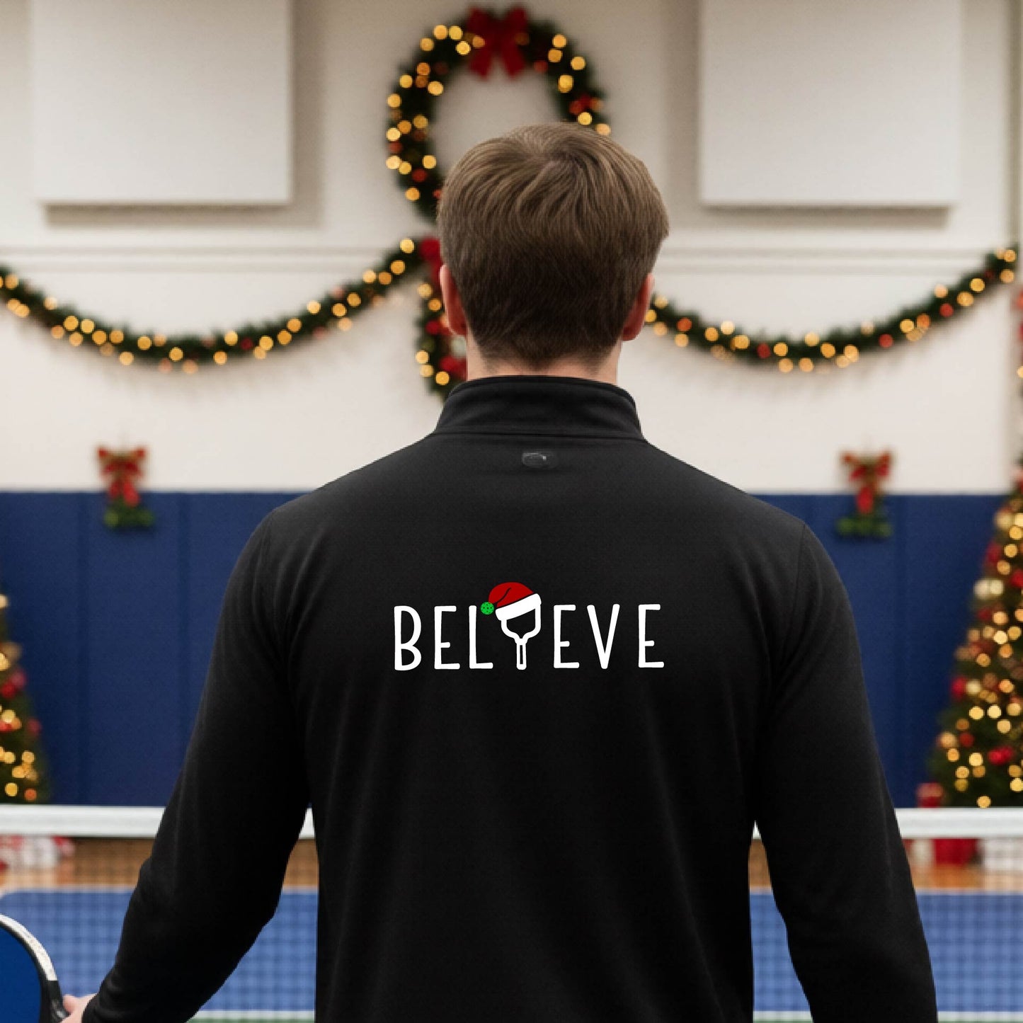 A smiling man wearing a black quarter zip long-sleeveshirt with the word 'BELIEVE' with the I repalced with a pickleball paddle wearing a Santa hat graphic stands on a pickleball court with a Christmas tree iand lights n the background.
