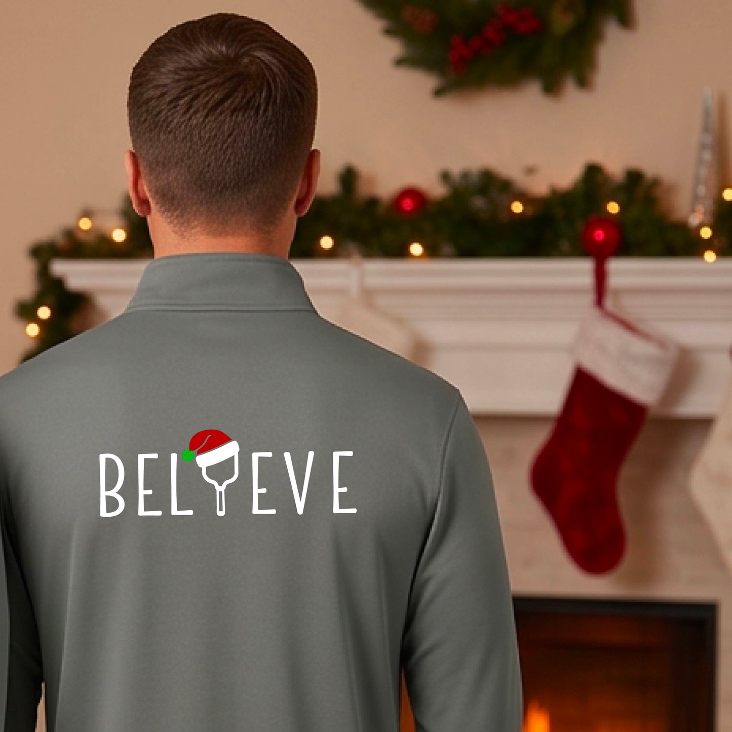 A smiling man wearing a gray quarter zip long-sleeveshirt with the word 'BELIEVE' with the I repalced with a pickleball paddle wearing a Santa hat graphic stands on a pickleball court with a Christmas tree iand lights n the background.