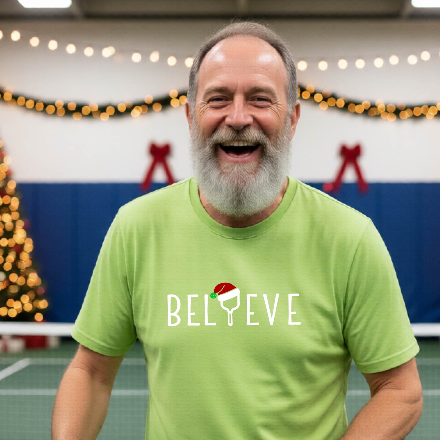 A man wearing a lime green short-sleeve t-shirt with a  pickleball graphic with text "BELIEVE" featuring a the letter I replaced with a pickleball paddle wearing a Santa hat.  