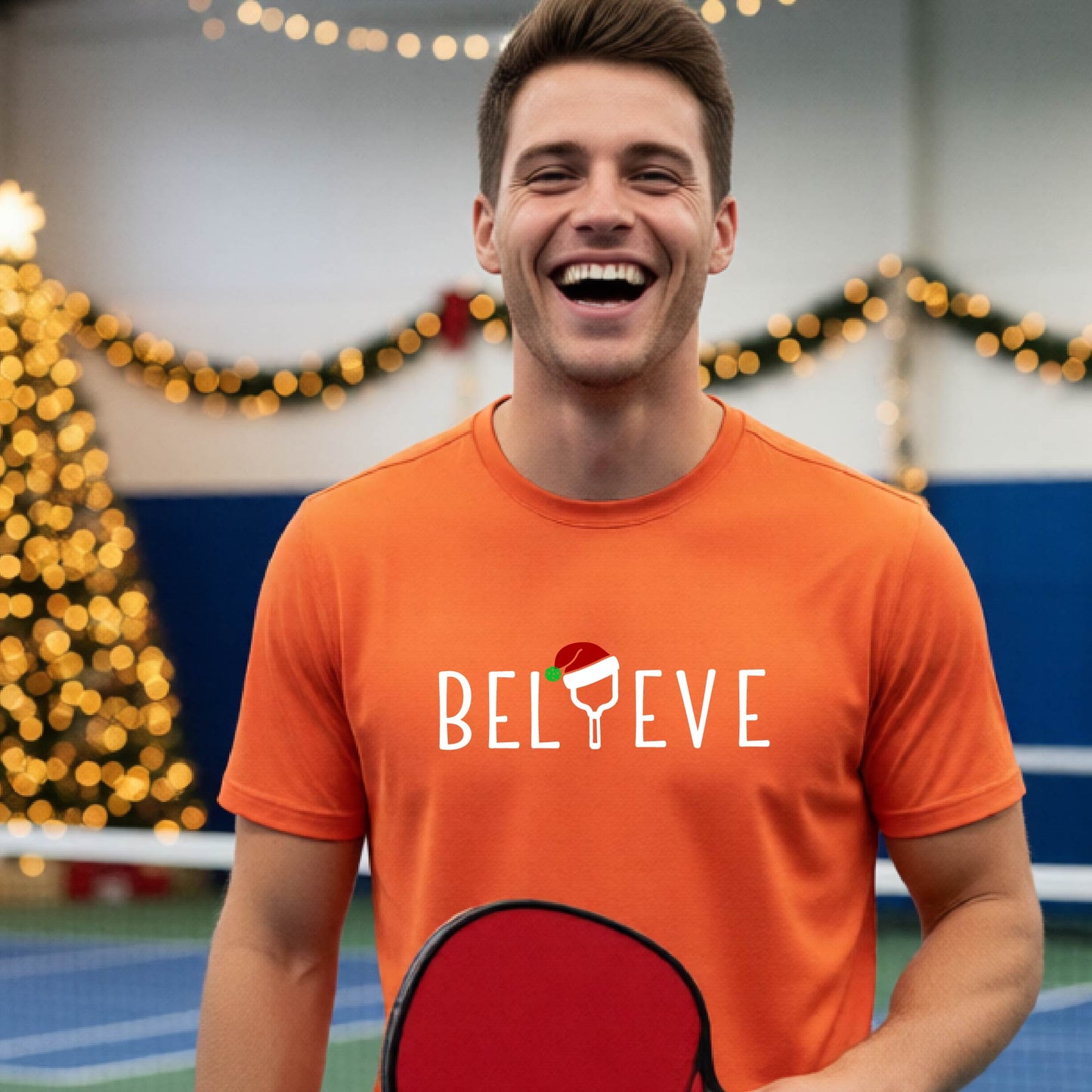 A man wearing an orange short-sleeve t-shirt with a  pickleball graphic with text "BELIEVE" featuring a the letter I replaced with a pickleball paddle wearing a Santa hat.  